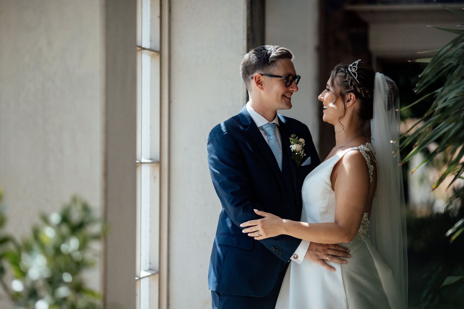 First look moment between couple in formal attire by sunlit window with tropical plants.