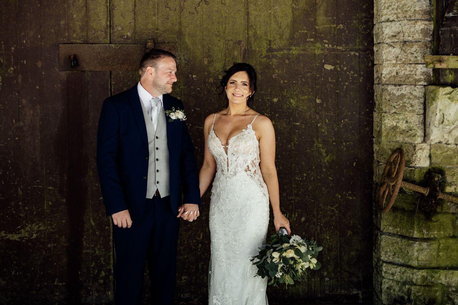 Wedding couple poses against rustic stone wall backdrop in formal attire.