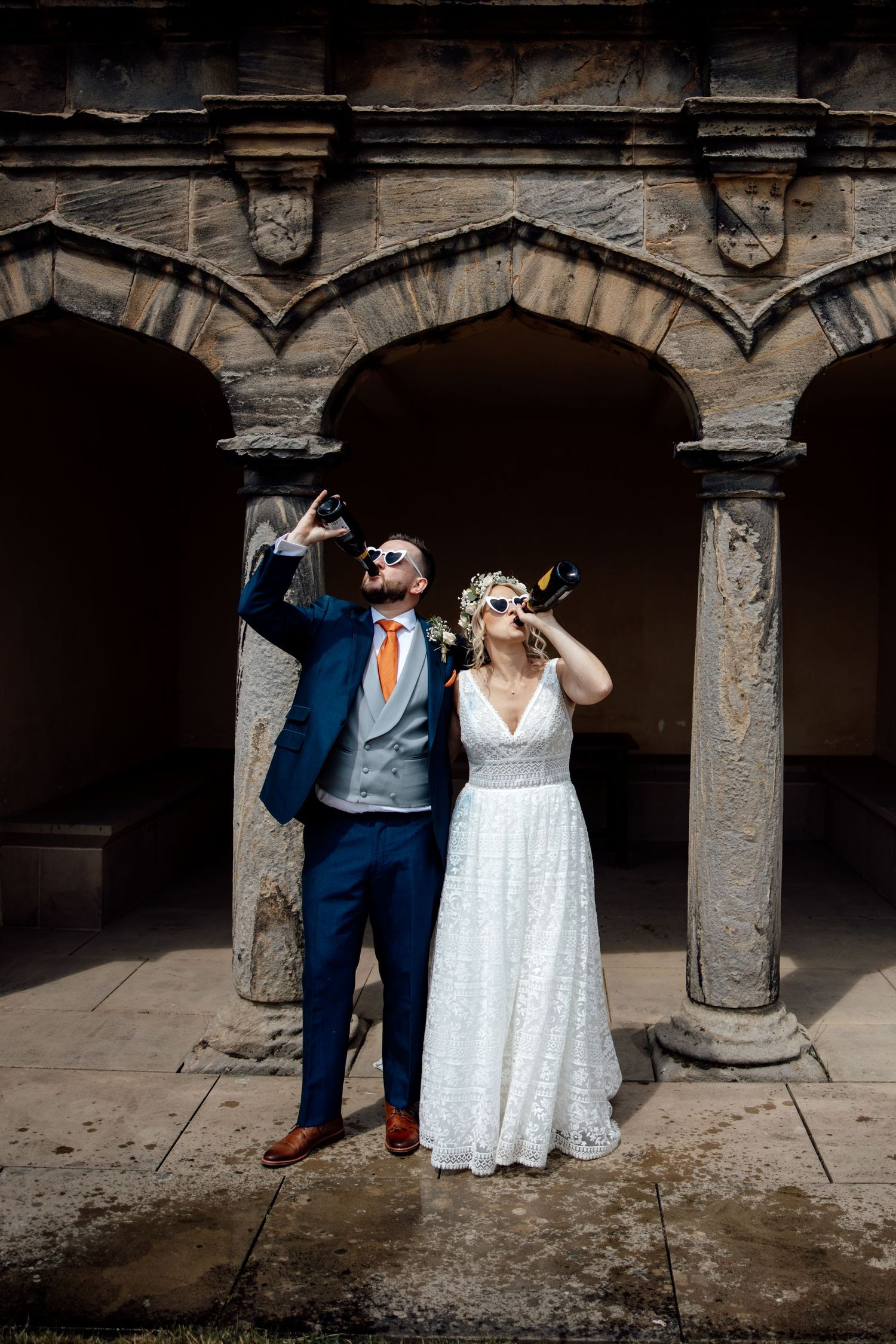 A couple poses playfully under stone archways in formal wedding attire.