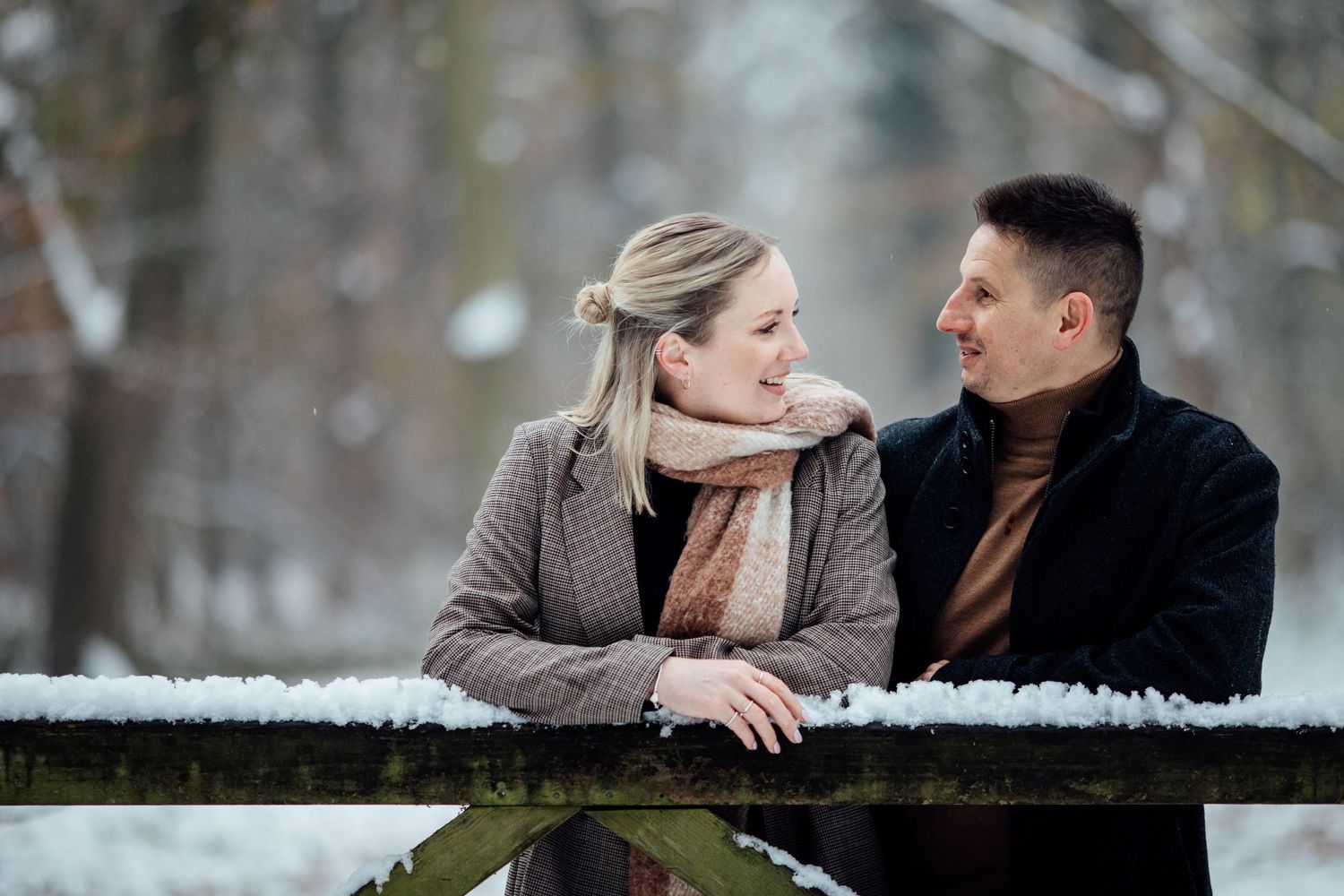 Intimate moment between couple leaning on wooden railing in snowy winter setting.