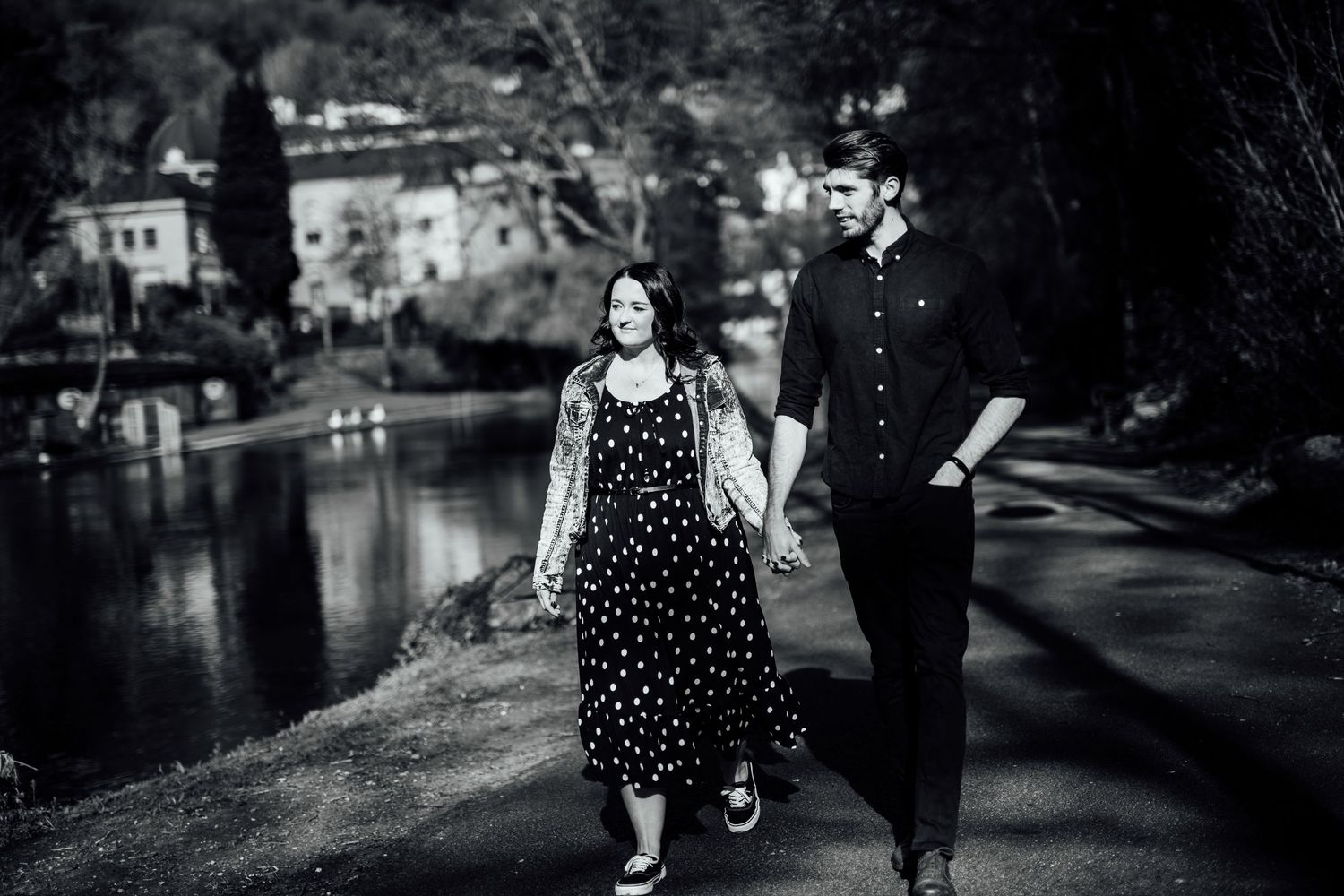 Two people stroll across an arched bridge over a tranquil pond in a park setting.