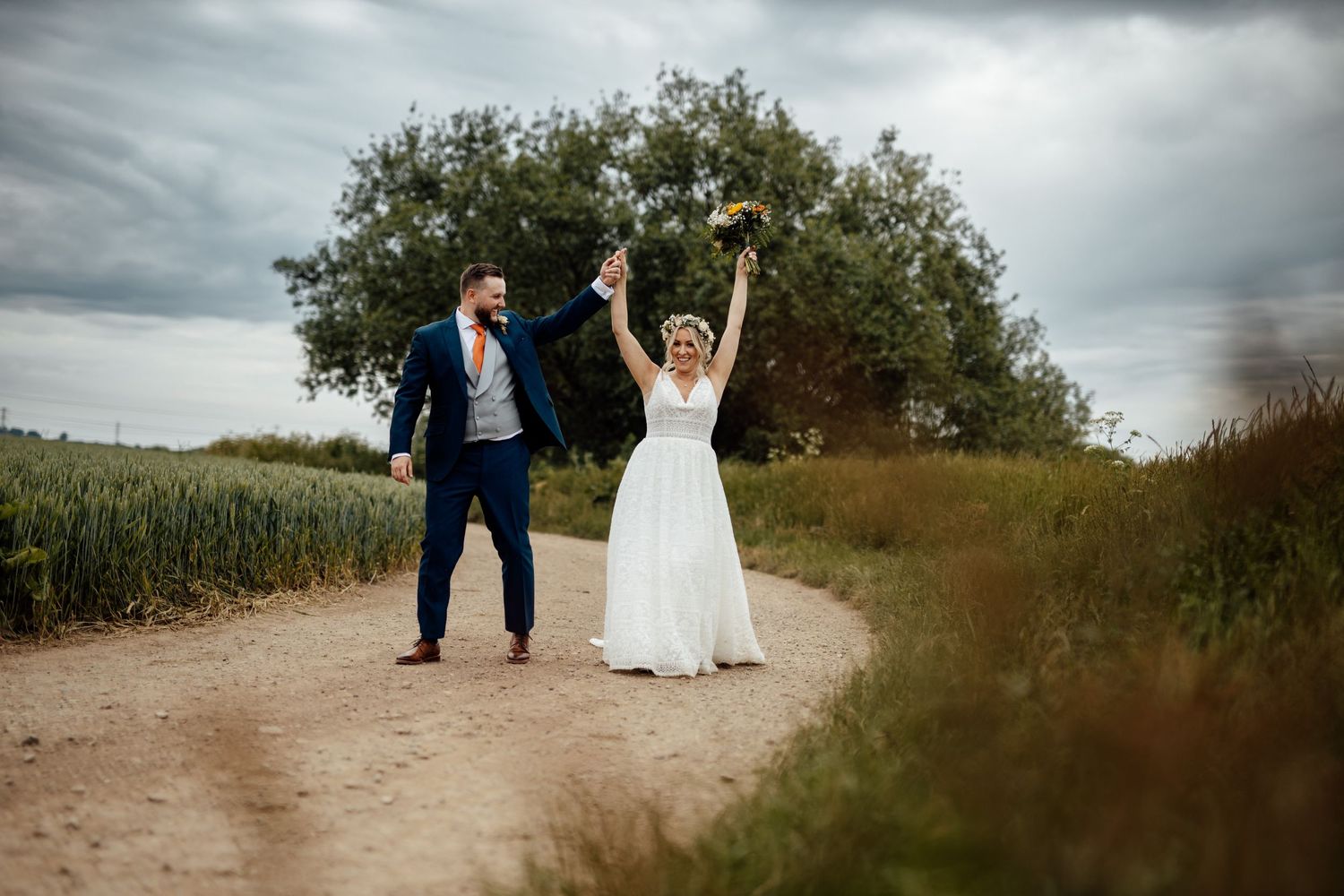 Dramatic wedding portrait with couple walking along rural path under moody sky.