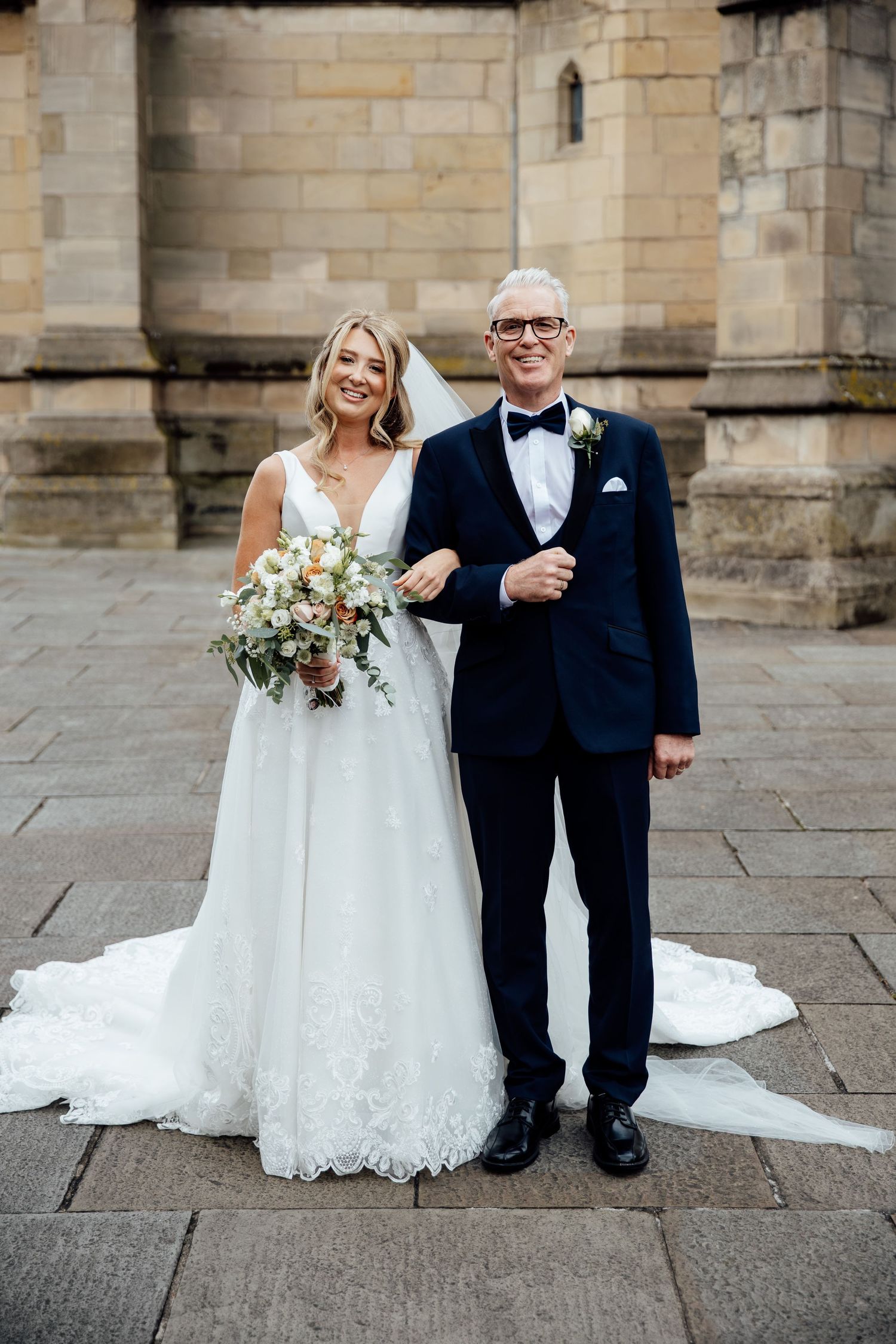 Bride poses with companion for formal photos outside historic stone church.