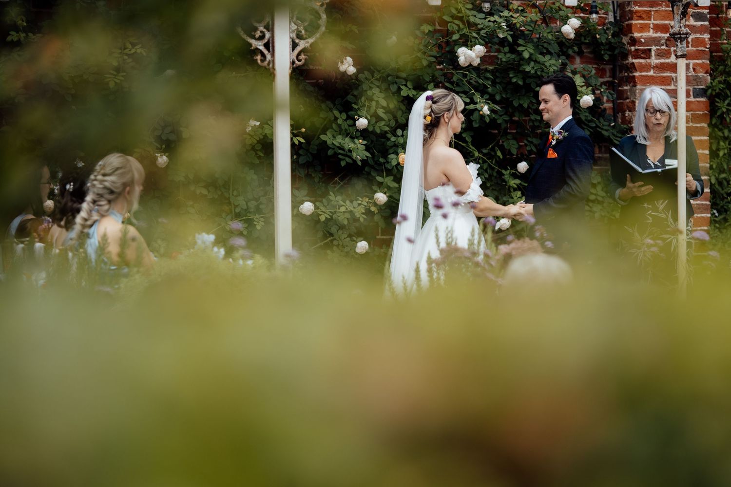 A wedding ceremony captured through foliage with a brick building in the background.