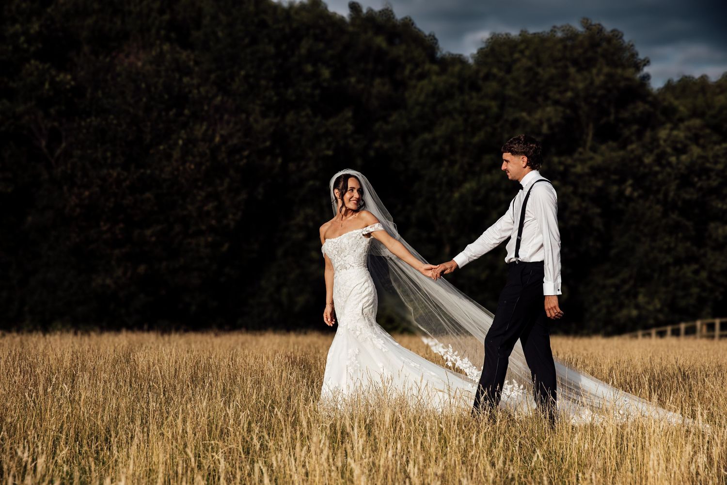 A couple walks through a golden wheat field at sunset with dramatic clouds overhead.