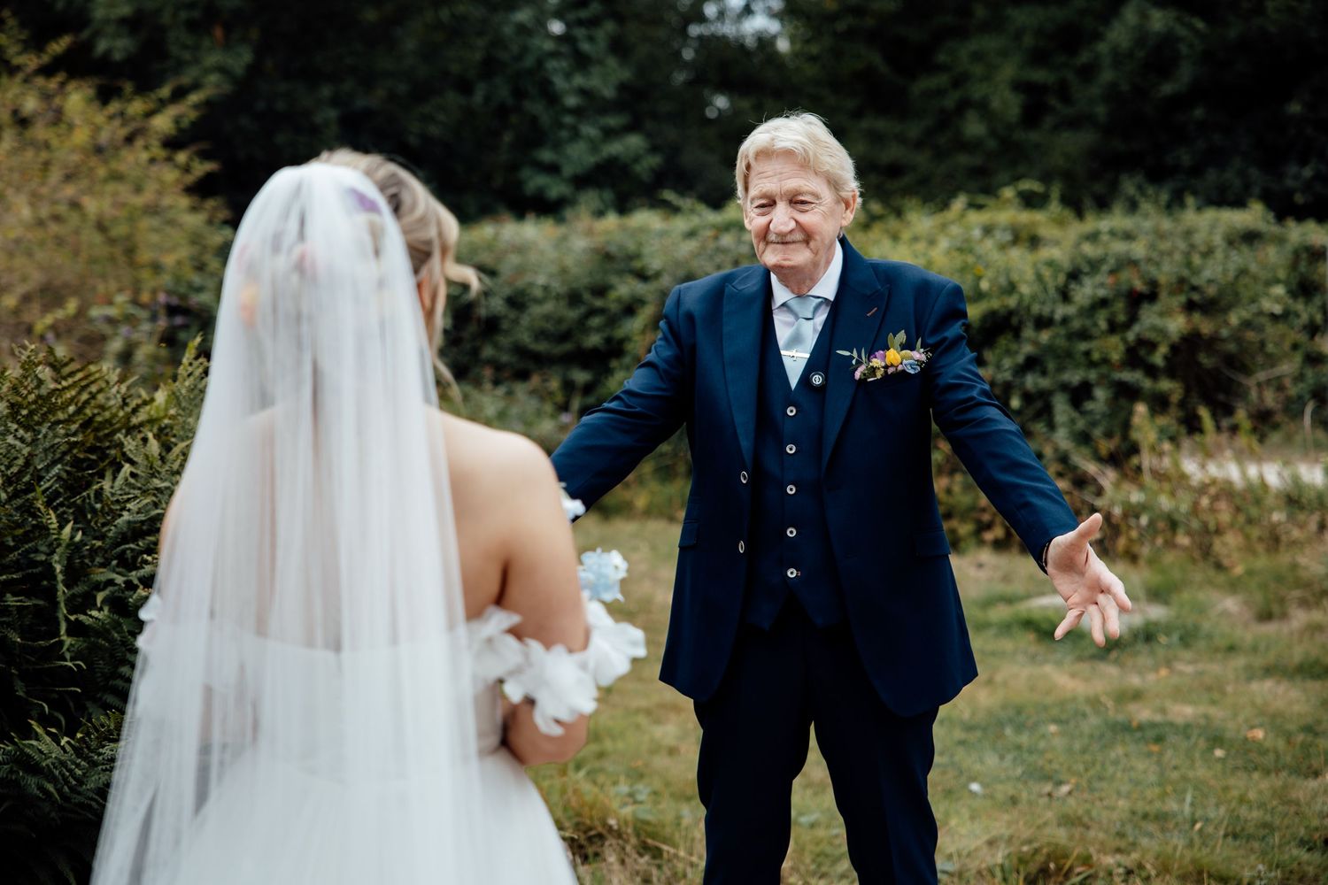 A heartwarming moment between a bride in white dress and veil with an elderly guest in a navy suit outdoors.