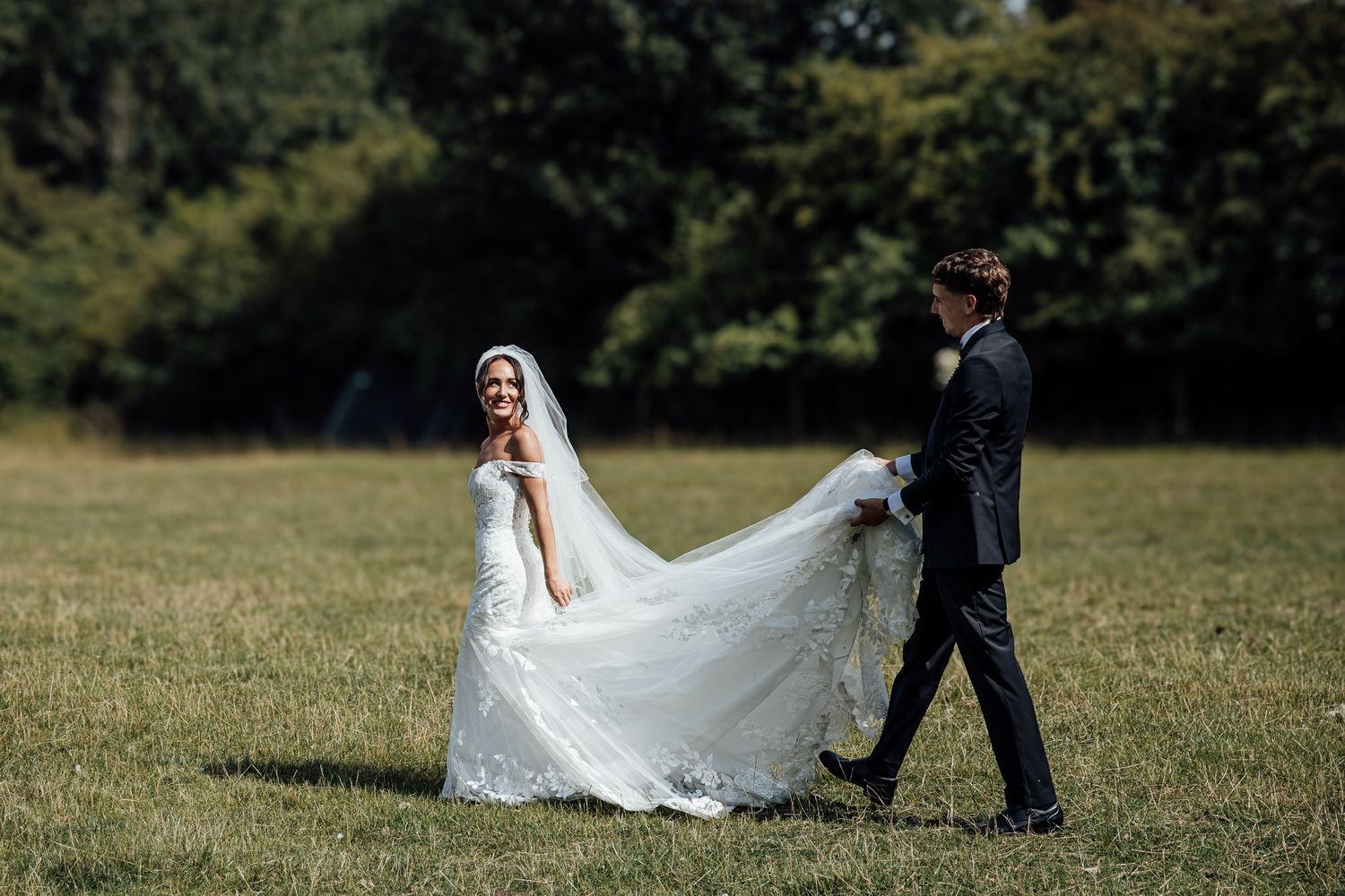 A couple poses in a field as the bride's long veil flows dramatically in the wind.