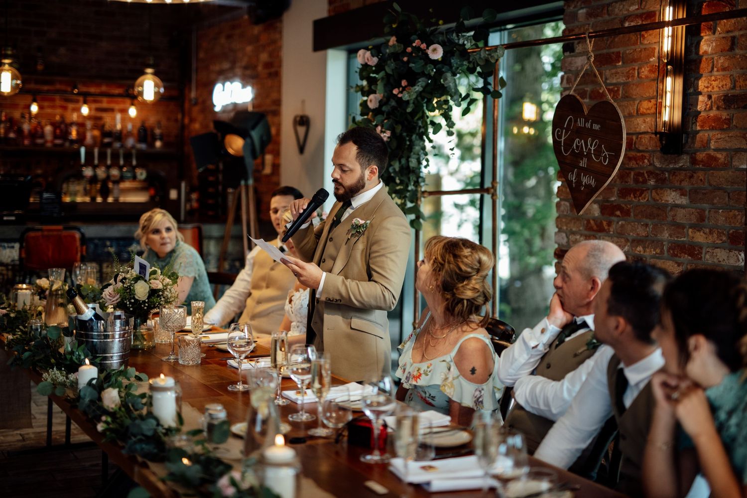 Wedding reception guests enjoying dinner at rustic tables with exposed brick wall backdrop.