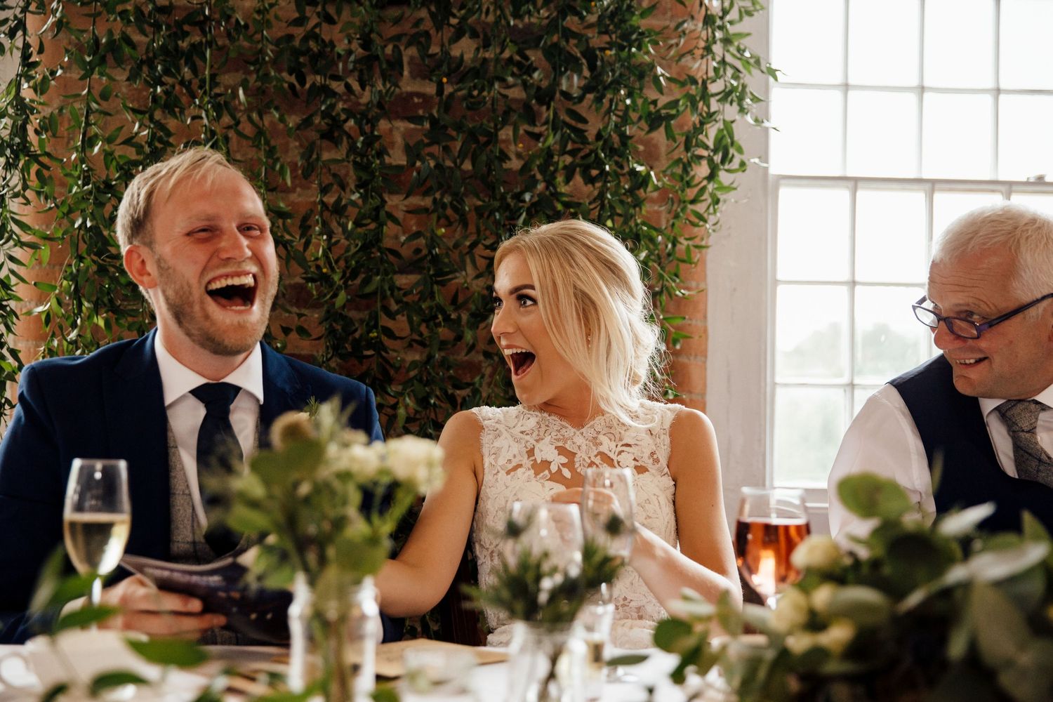 Couple shares a joyful laugh during their wedding reception dinner surrounded by greenery and champagne glasses.