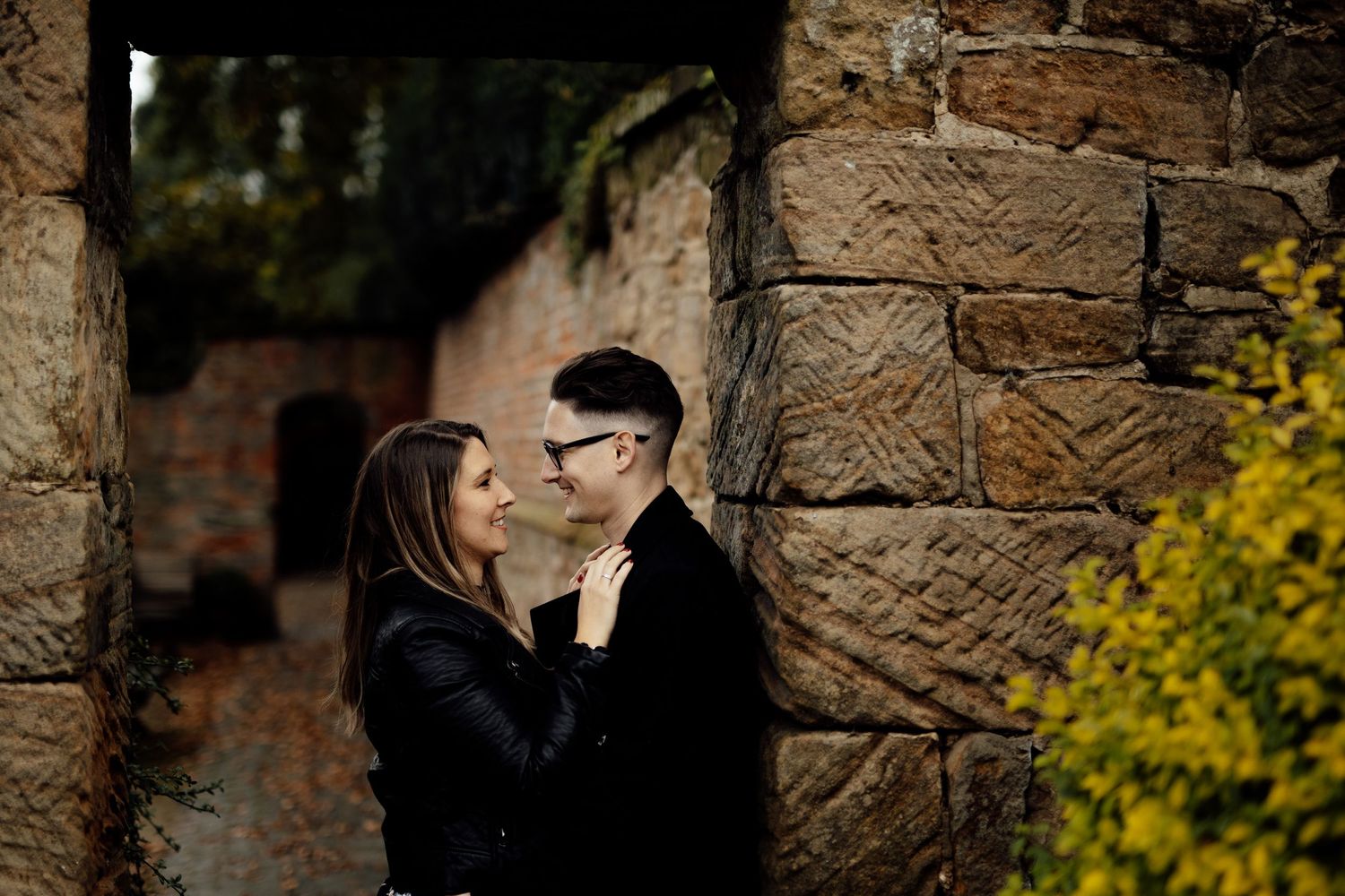 A couple poses near stone walls with yellow foliage creating a romantic autumn atmosphere.