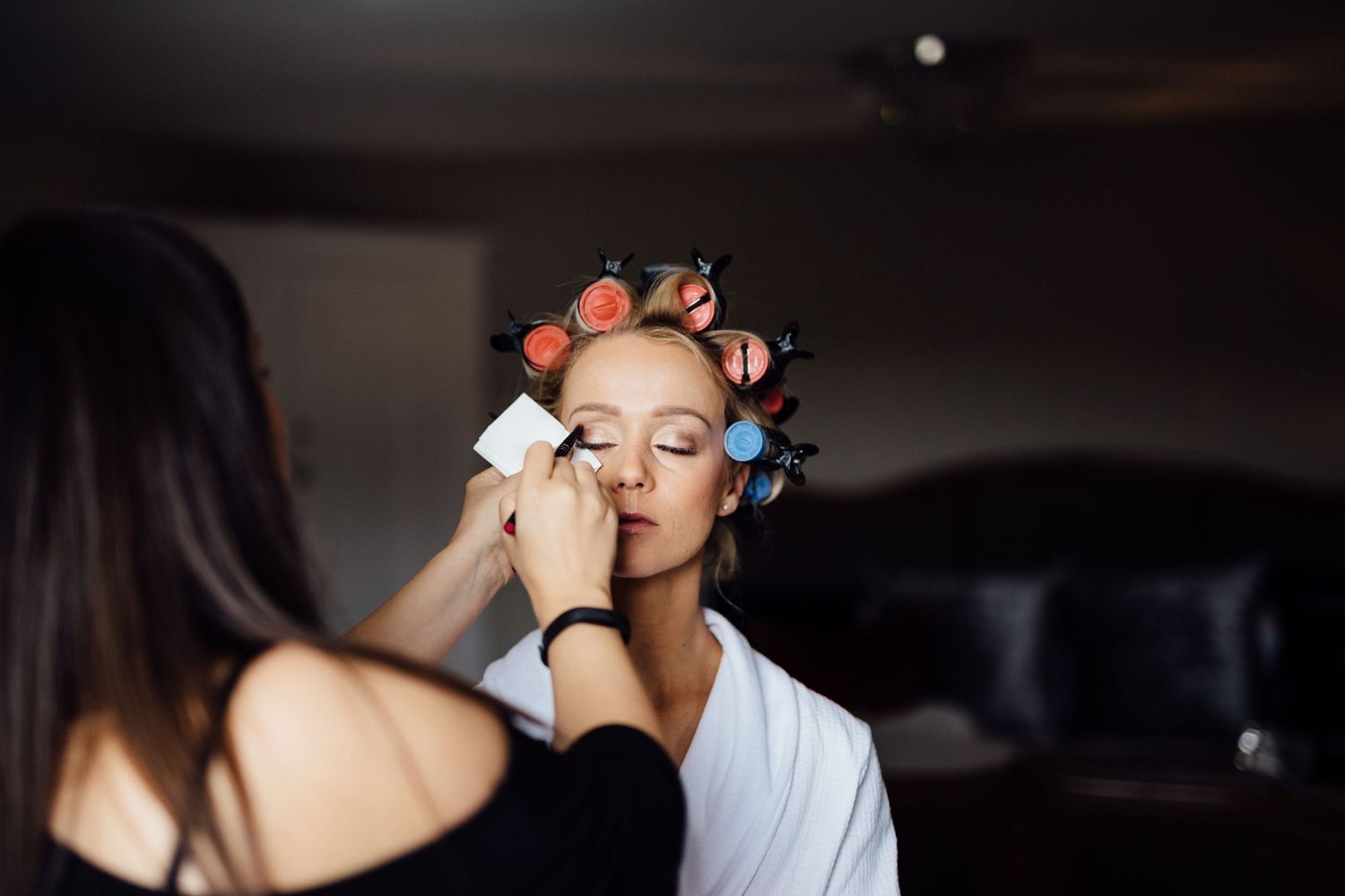 Getting ready scene with hair rollers and makeup application in a dim room.