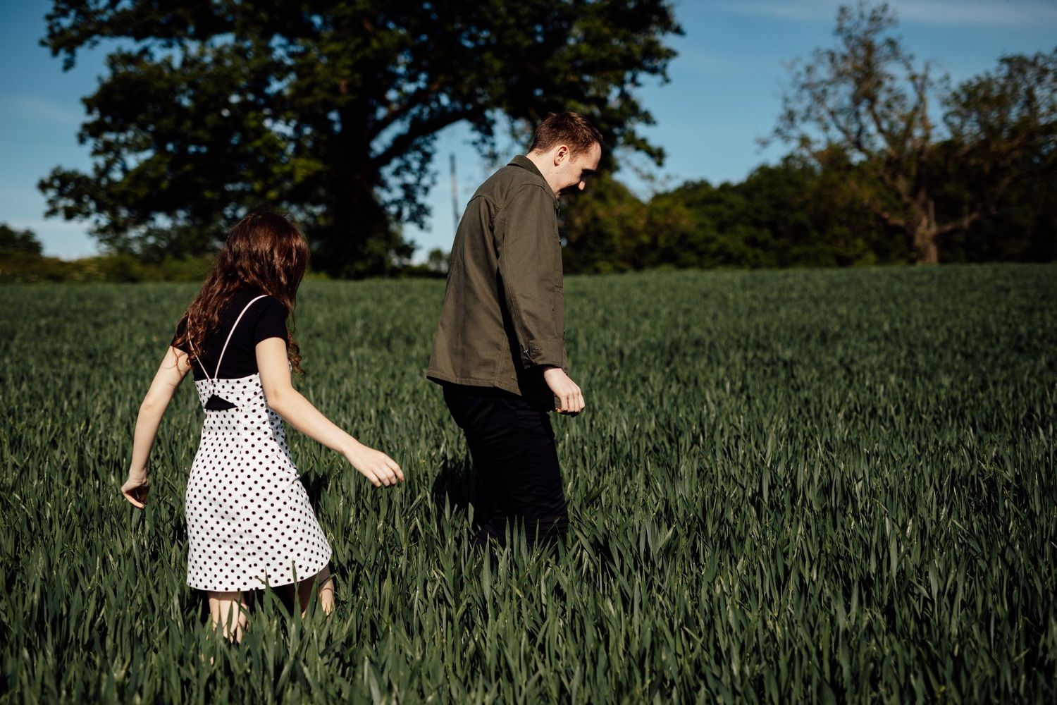 Two people walking through tall green wheat fields on a bright afternoon.