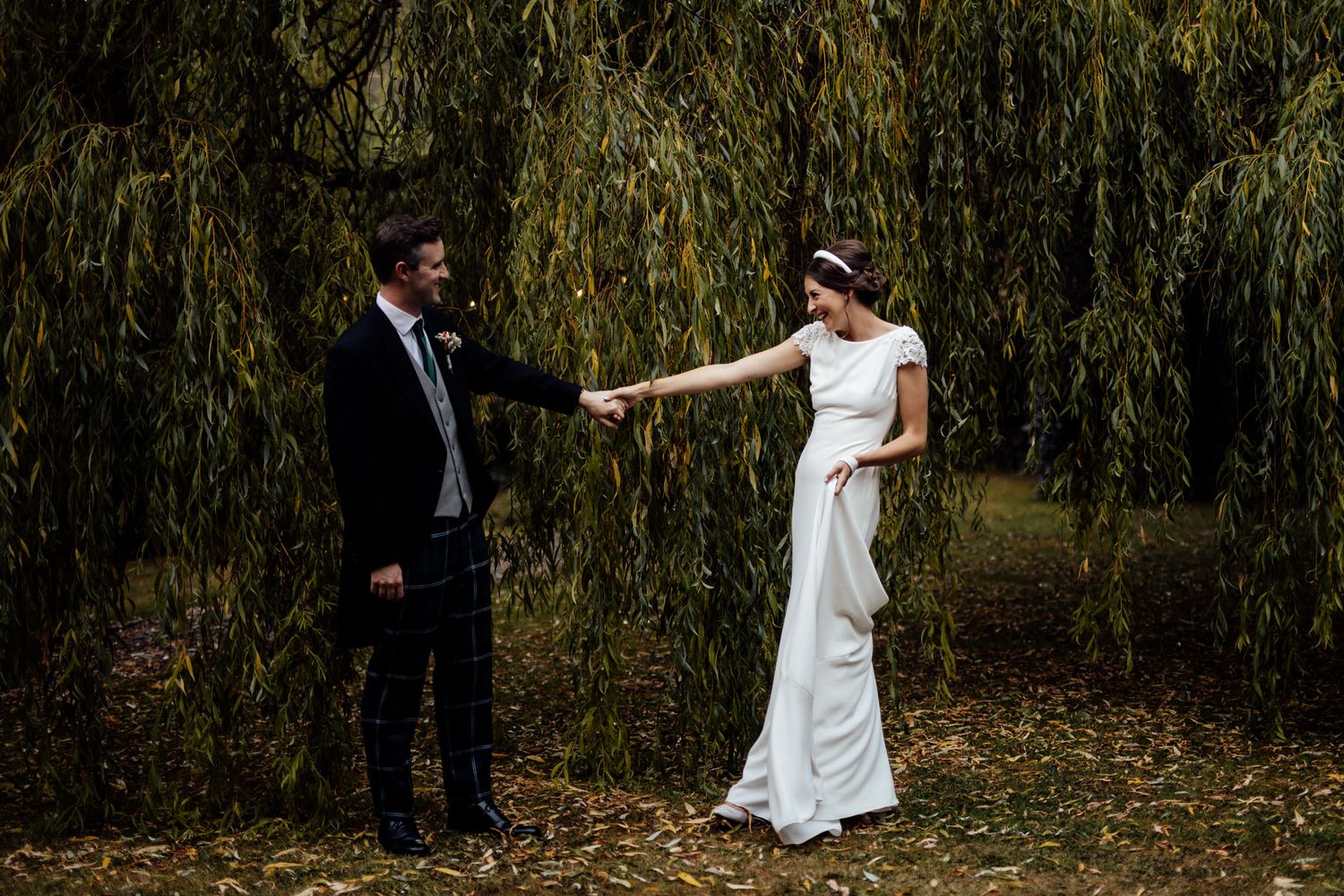 Wedding party posing for photos near weeping willow trees in a park.
