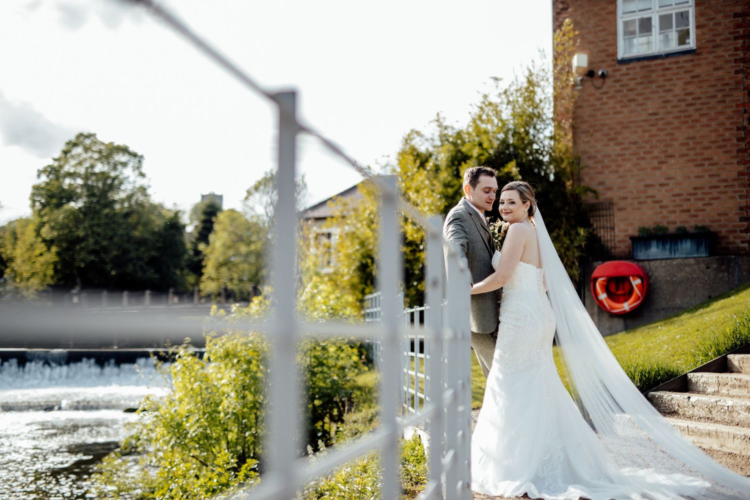 Wedding photos taken along a waterfront with brick buildings and greenery in the background.
