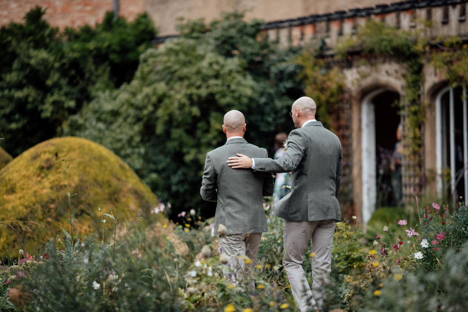 Two people walking through a manicured garden with stone architecture in the background.
