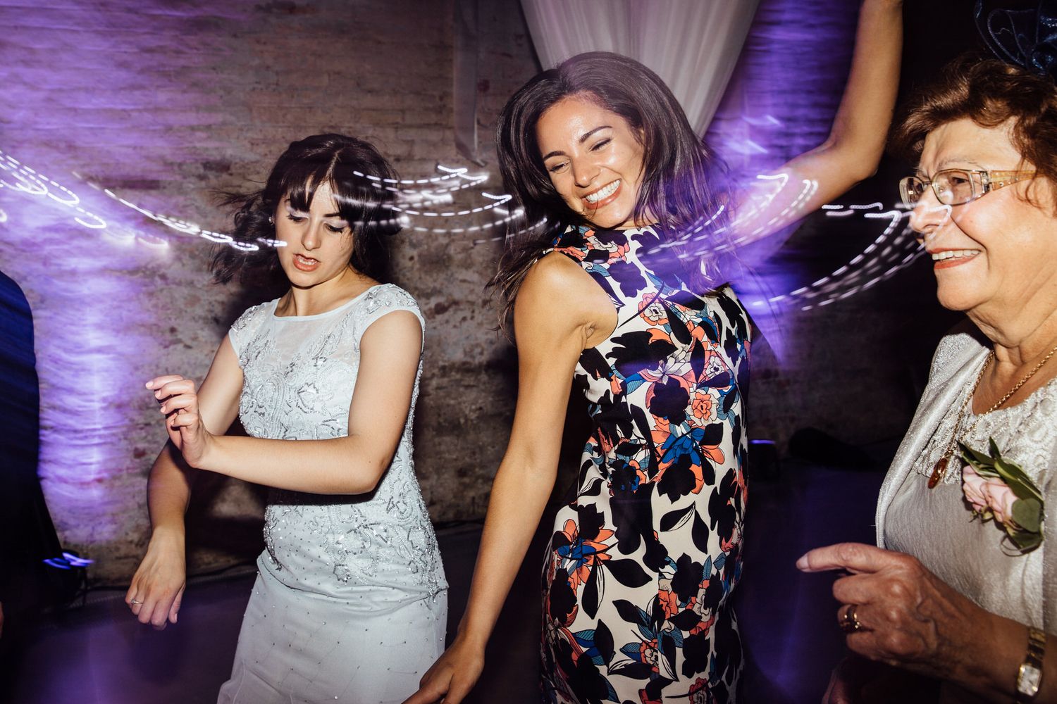 Wedding guests dancing and celebrating at a reception with purple lighting and white drapes.