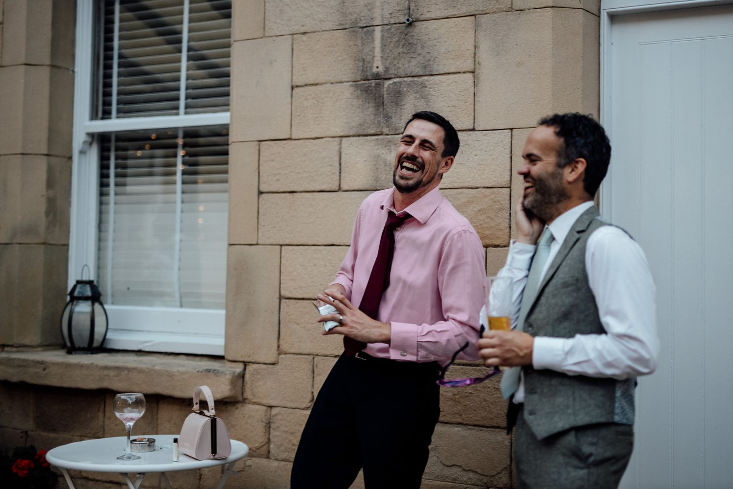 Wedding guests chatting and laughing outside a stone building on a sunny day.