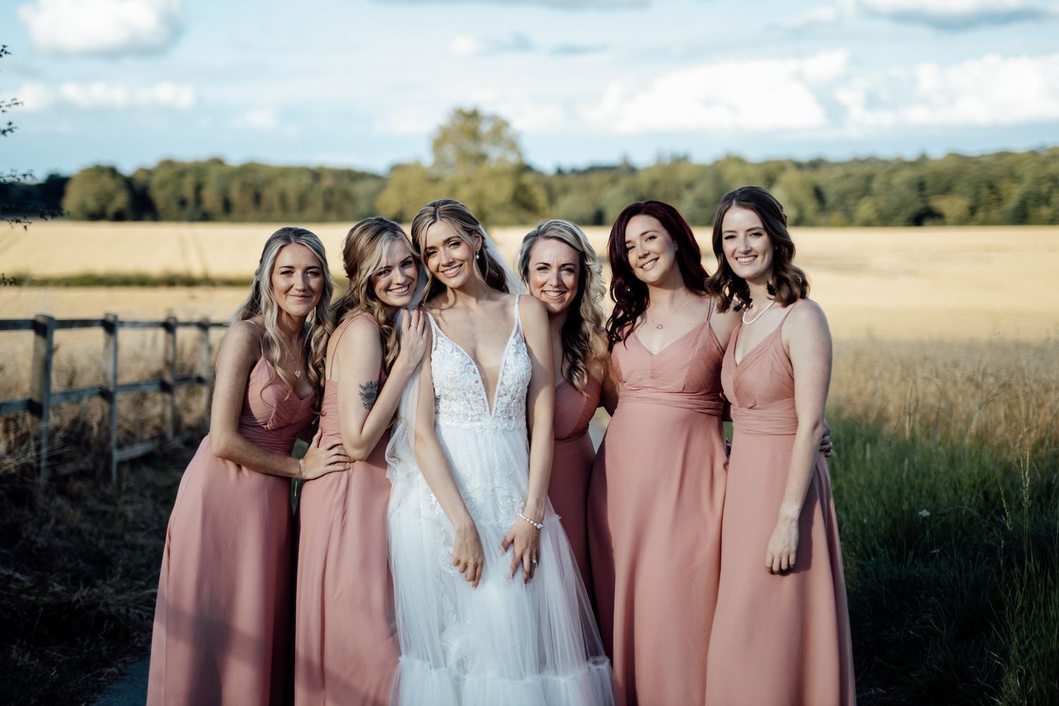 Bridal party in blush pink dresses gather close together on rustic path near wheat field.