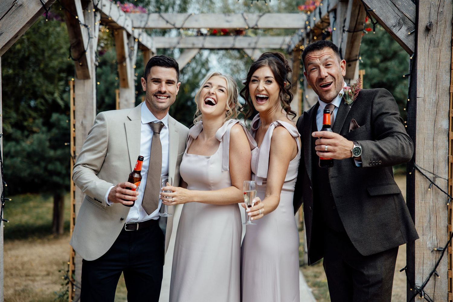Two bridesmaids in grey dresses laugh joyfully with groomsmen holding drinks at outdoor wedding.