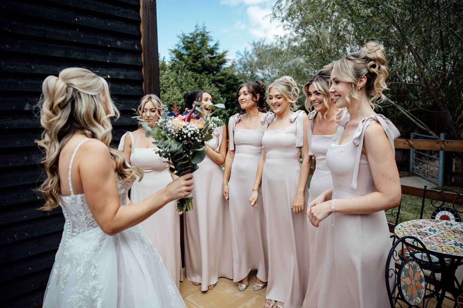 A bride in a white gown shows her bouquet to smiling bridesmaids in blush pink dresses outdoors.