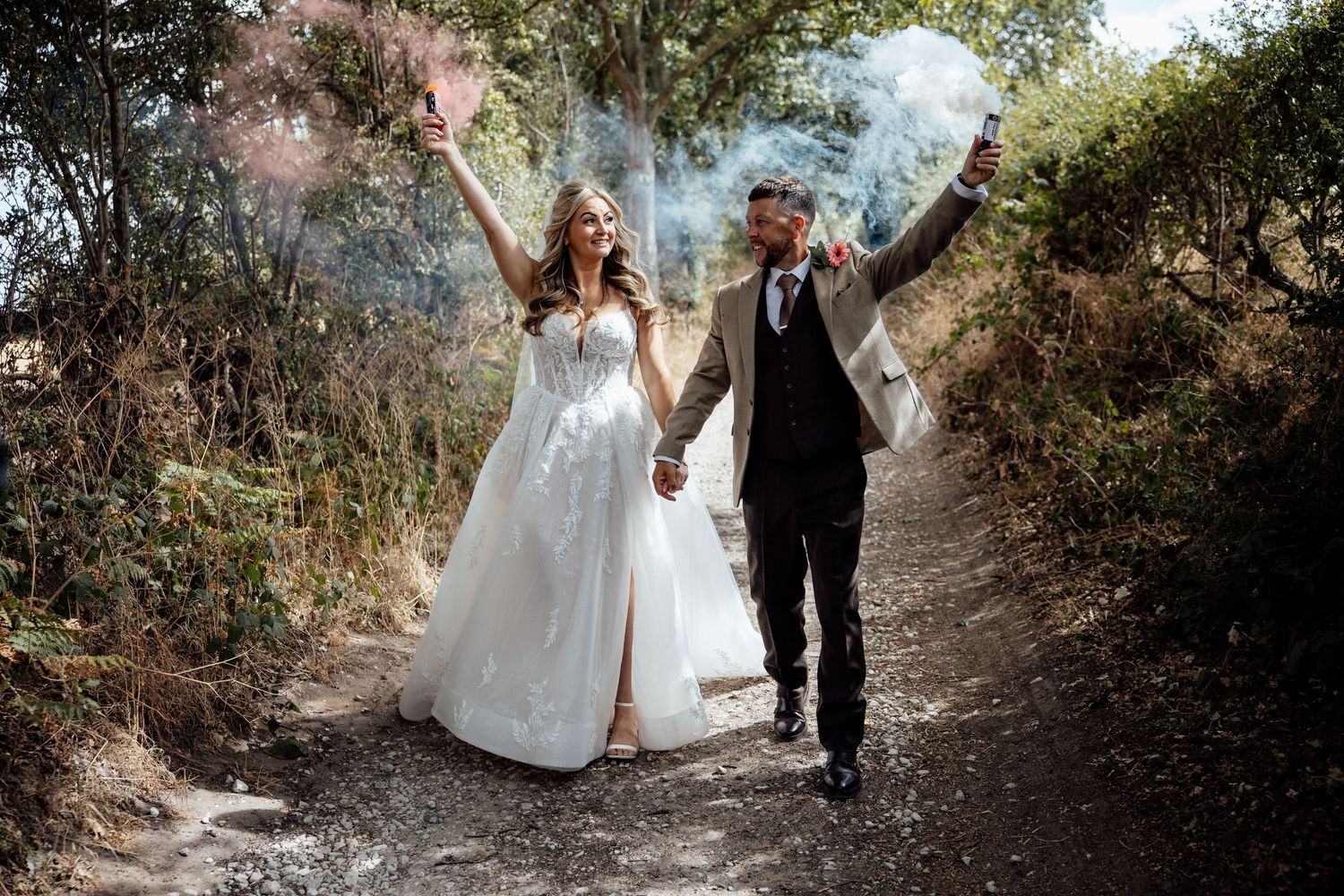 Joyful newlyweds holding colorful smoke bombs while walking along a woodland path after wedding.