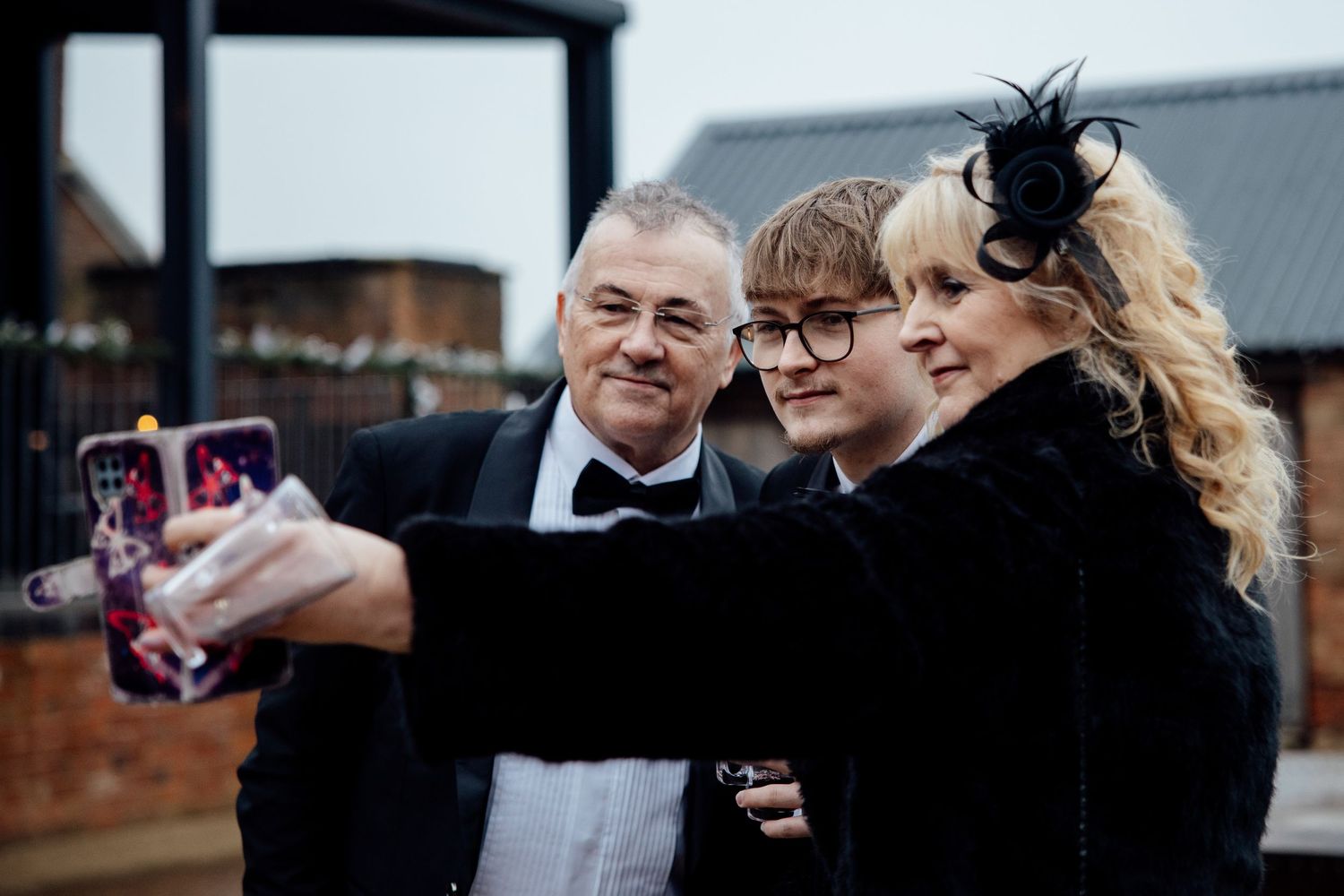 Wedding guests in formal attire take a group selfie outdoors.