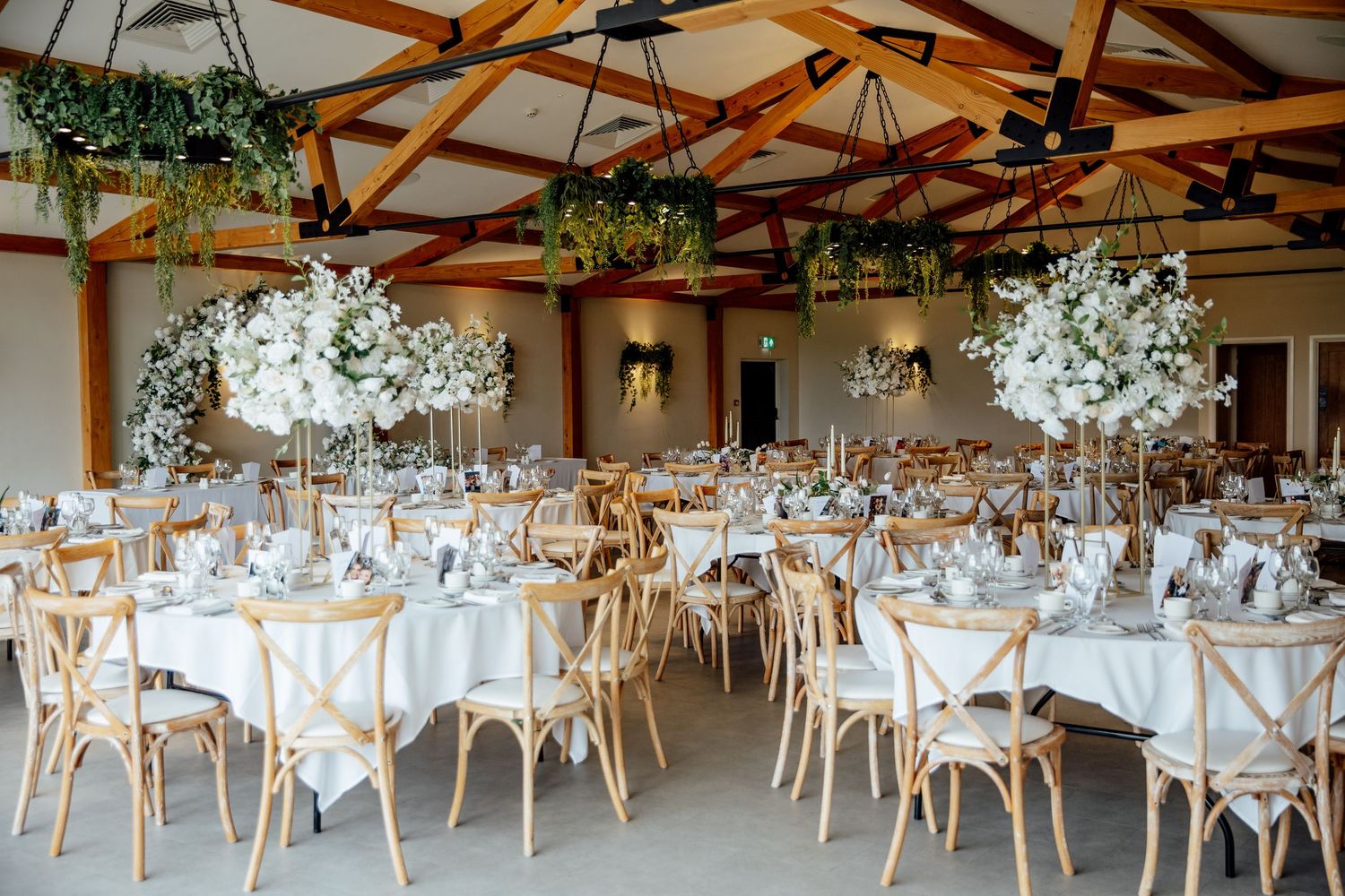 Elegant wedding reception venue with white table settings, wooden crossback chairs and tall white floral centerpieces.