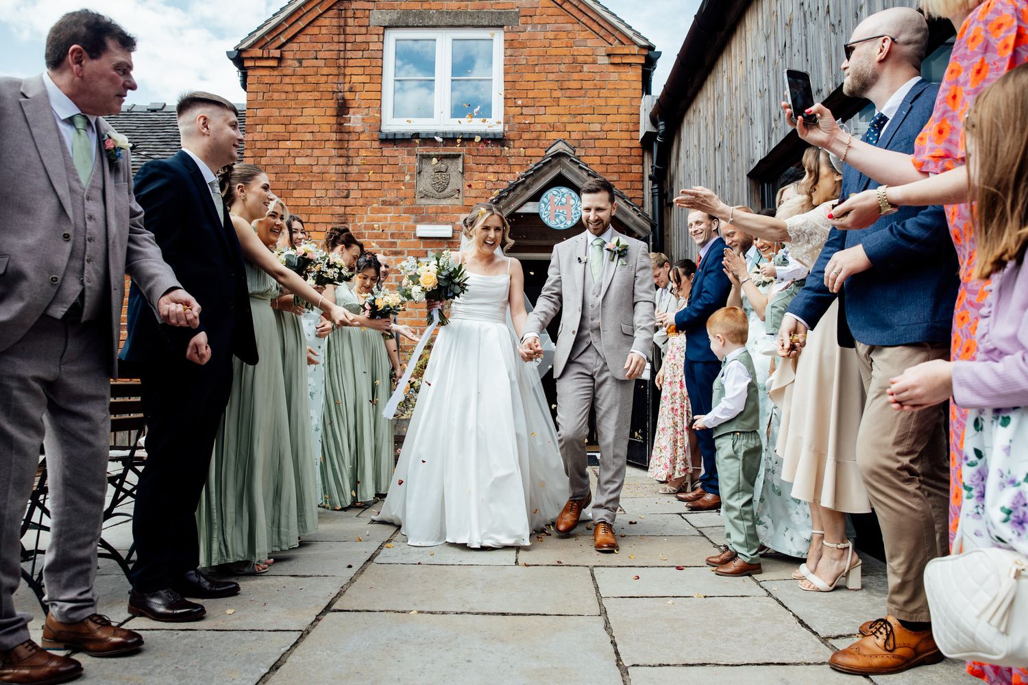 Wedding ceremony lineup in front of a rustic brick building with guests watching.