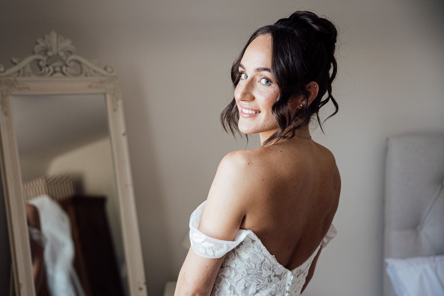 Bride in off-shoulder lace dress smiling over shoulder in elegant bridal preparation room.