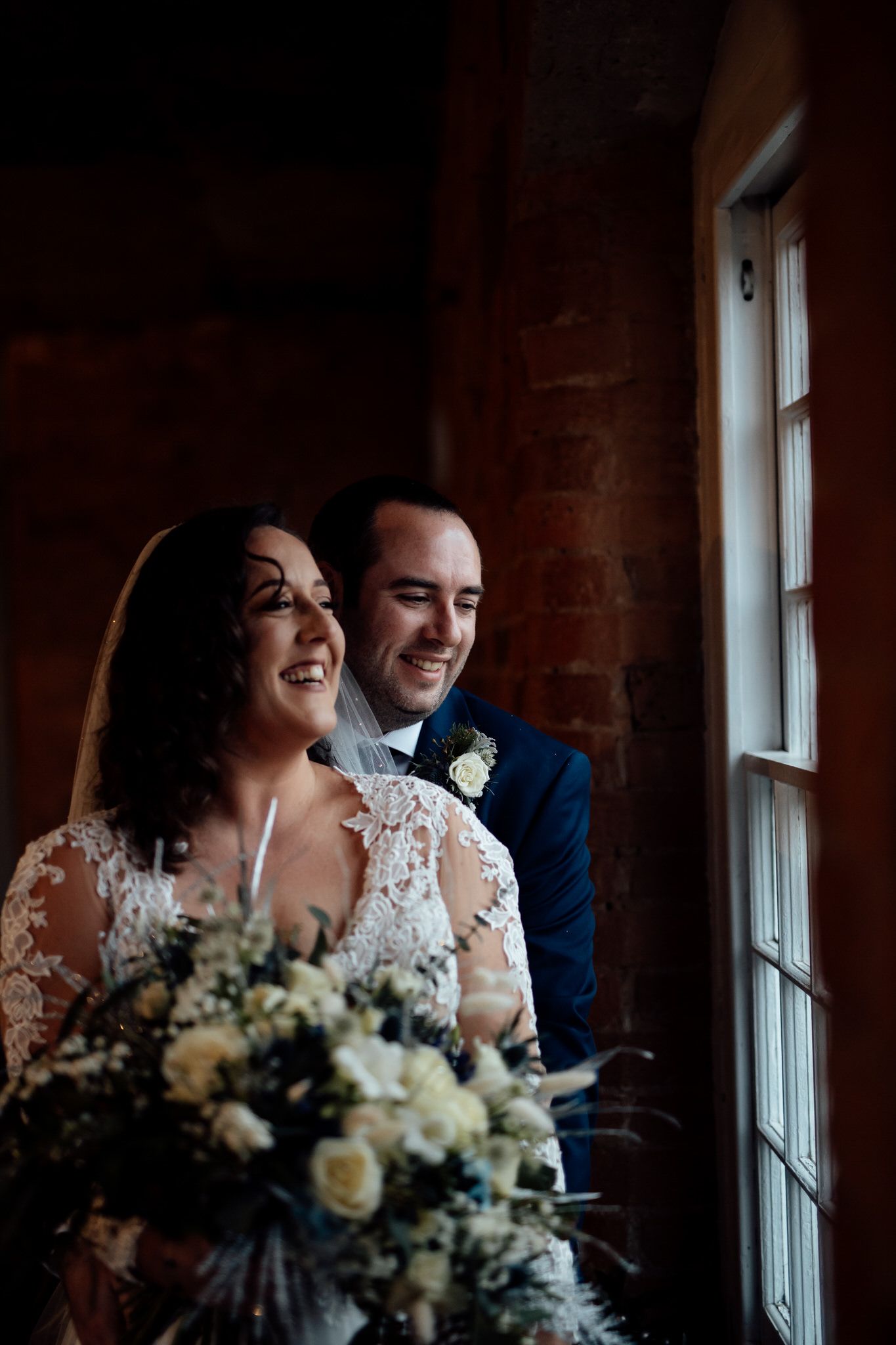 Romantic portrait of newlyweds by window with white floral bouquet in brick-walled room.