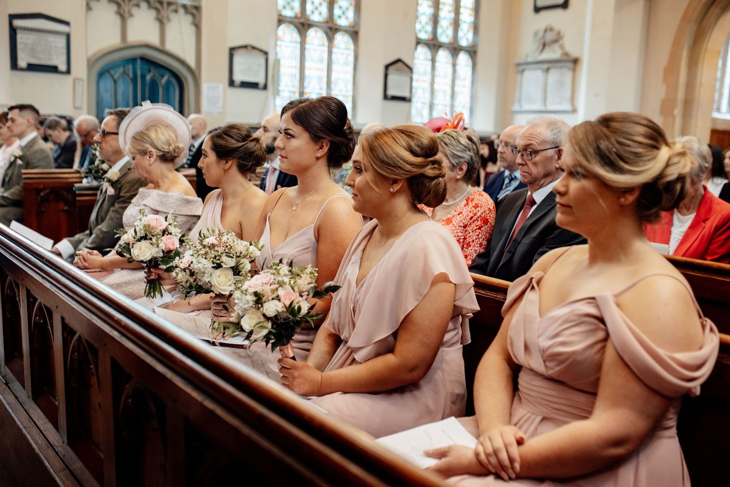 Bridesmaids in blush dresses seated in church pews holding floral bouquets.