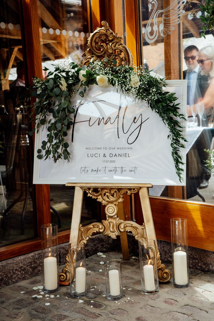 Elegant wedding welcome signs with greenery and candles displayed on gold easels.
