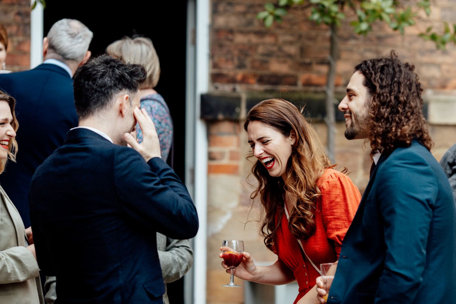 A group of wedding guests enjoying conversation and drinks at an outdoor reception with exposed brick walls.