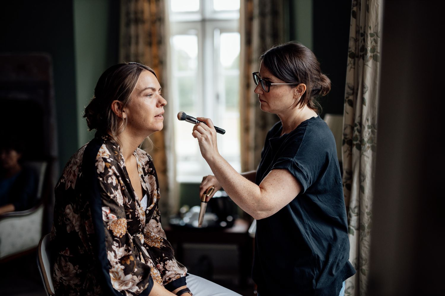 Professional makeup application in progress by window light in an elegant room.