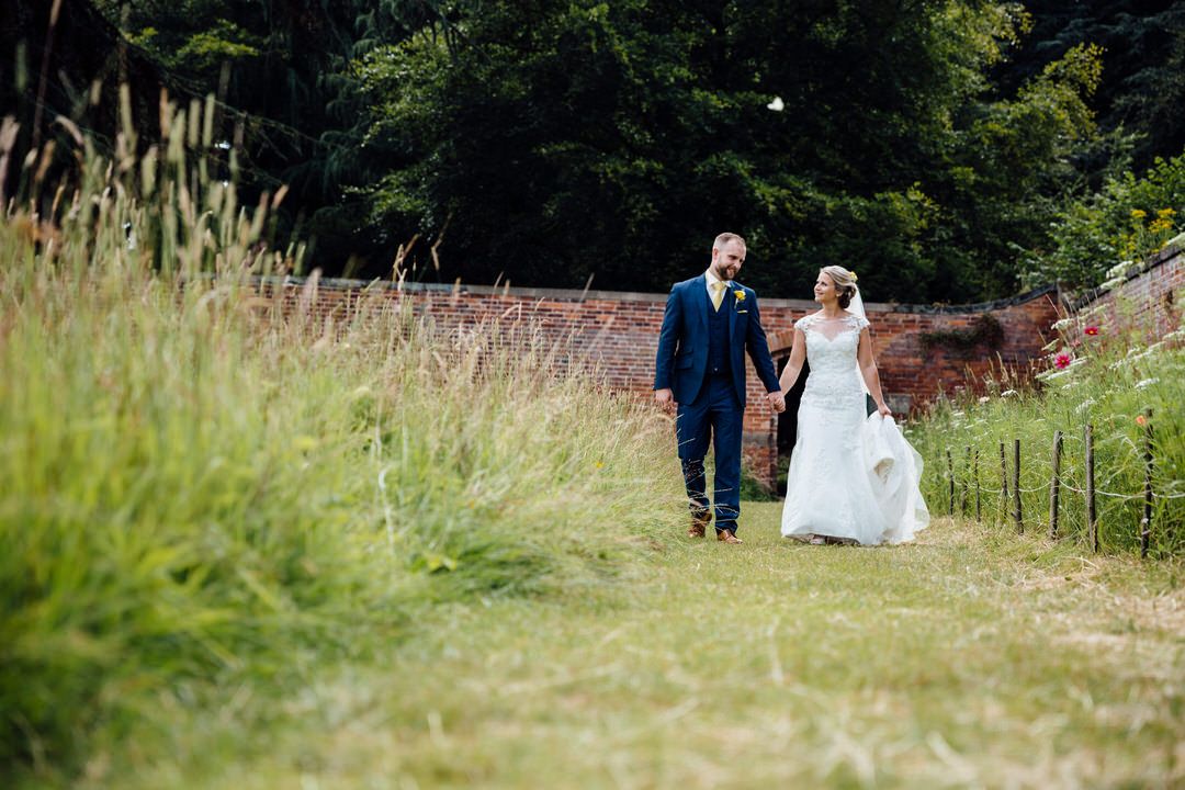 Newlyweds walking hand in hand through a scenic meadow path.