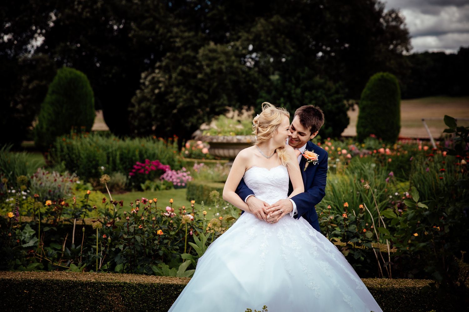 Newlyweds share intimate moment in blooming garden with dramatic sky.