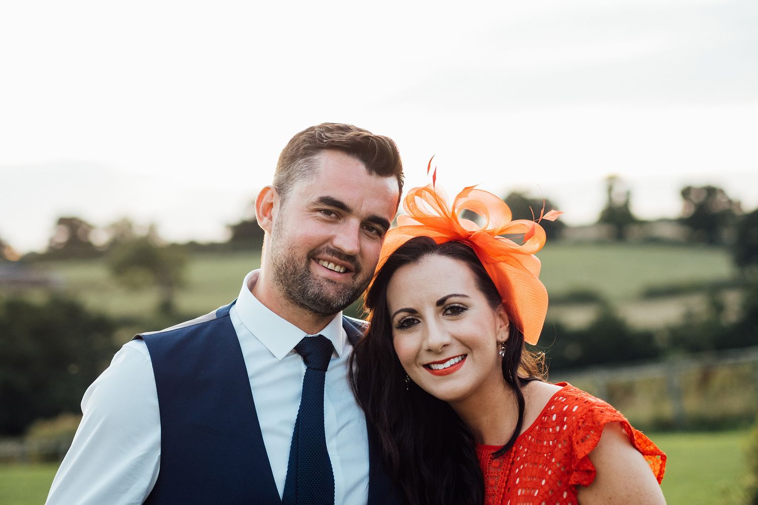 Wedding couple in formal attire with bright orange accessories in countryside setting.