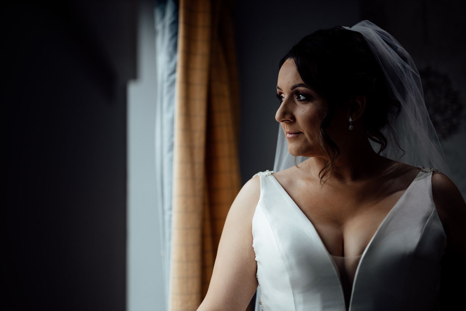 Dramatic window light illuminates a bride in a classic white wedding gown.