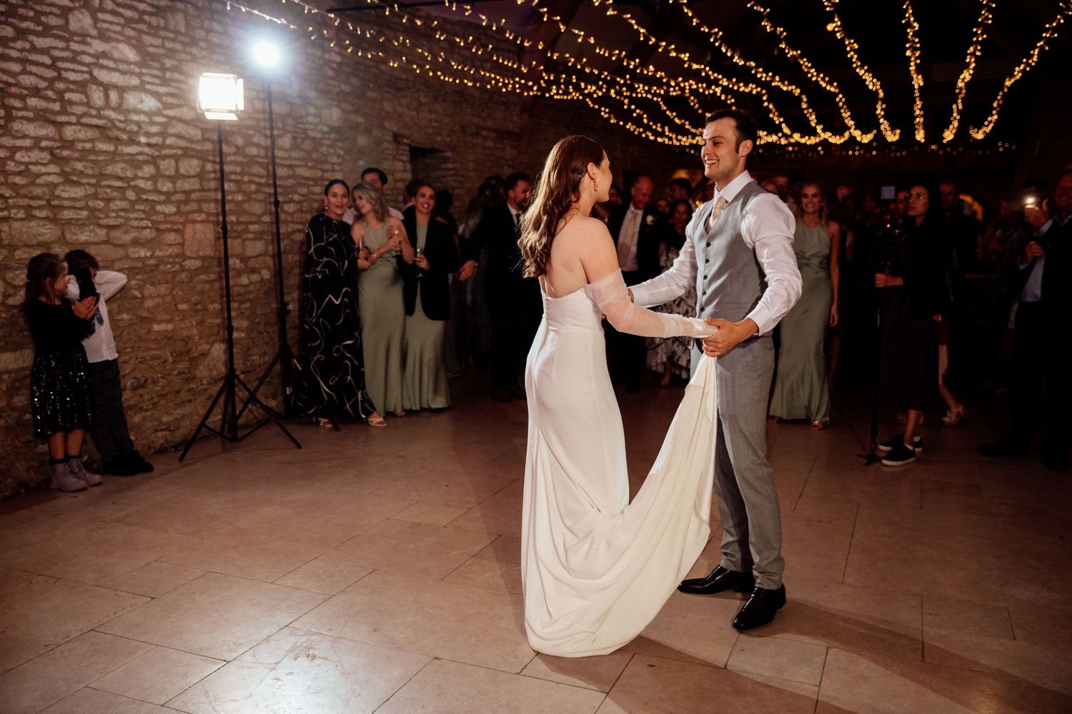 Couple sharing first dance under string lights in rustic brick-walled venue.