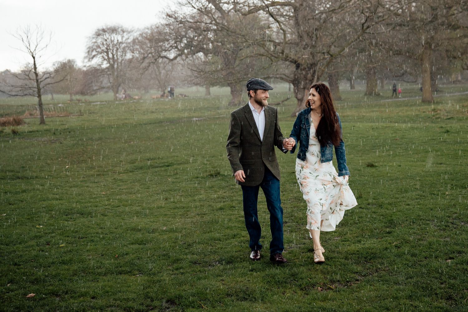 A couple strolls through a green field, with her dress flowing in the breeze as they share a joyful moment.