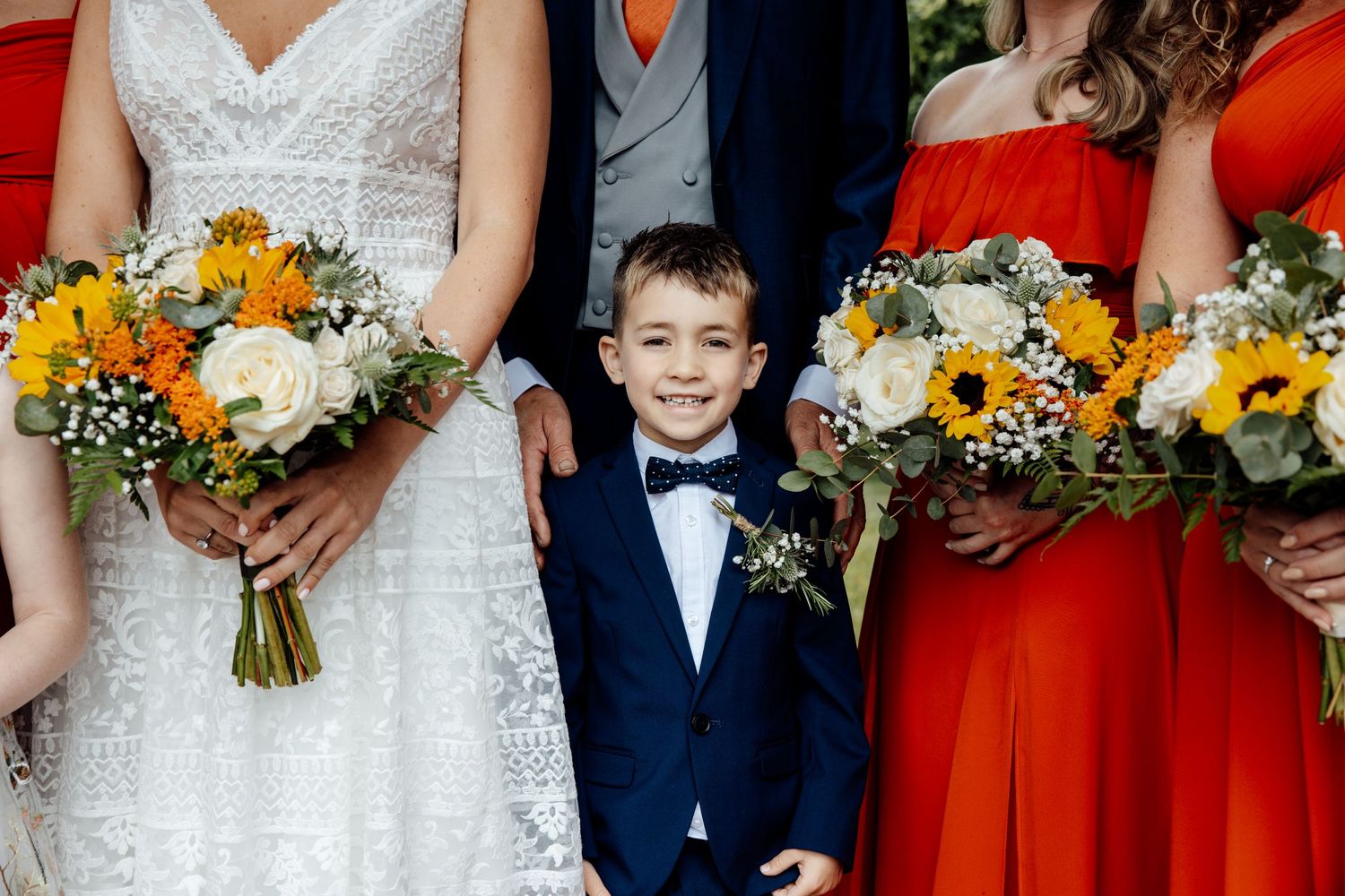 Wedding party in navy suits and coral dresses holding sunflower bouquets.