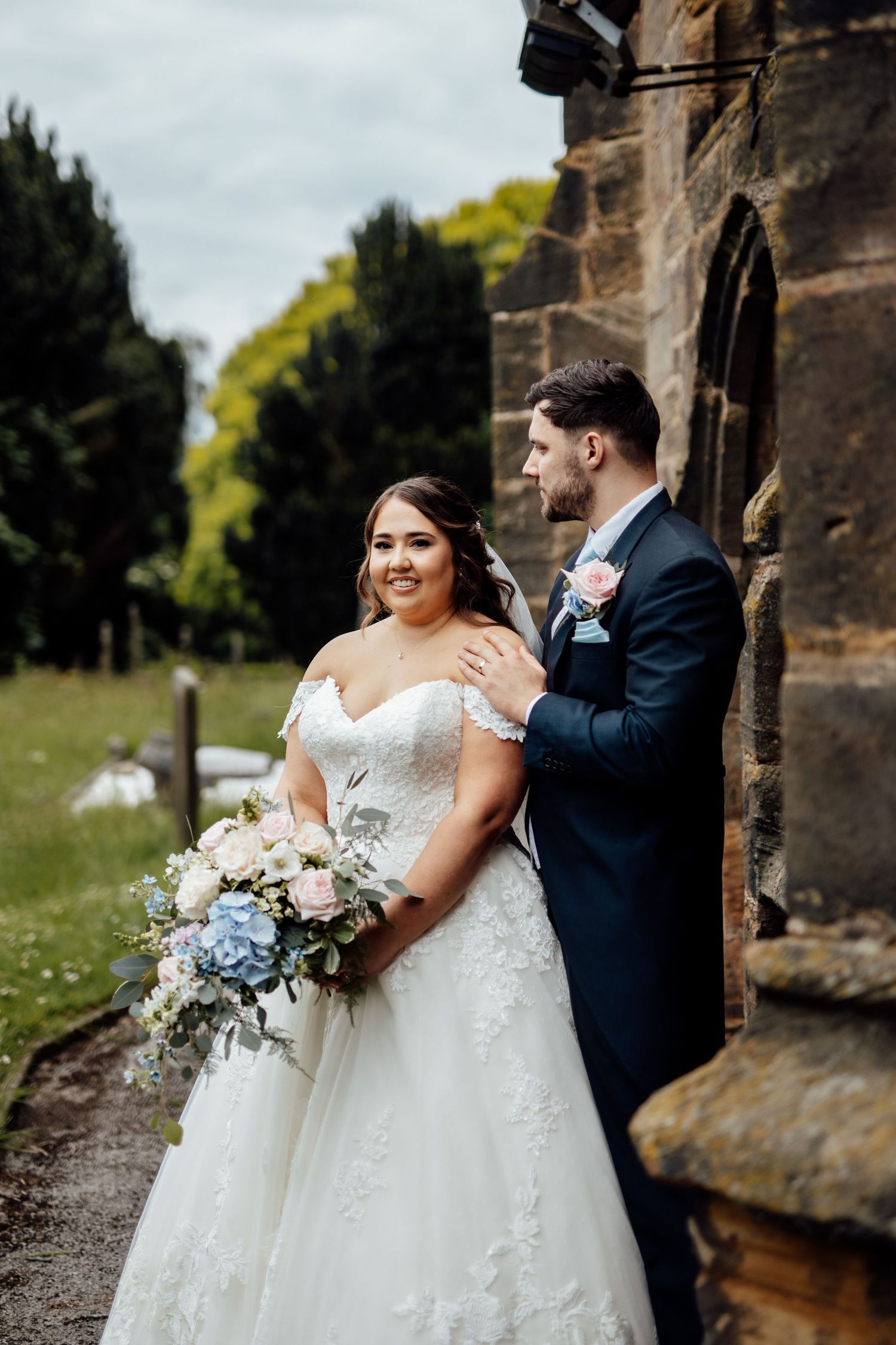 Couple in wedding attire posing against stone church exterior.