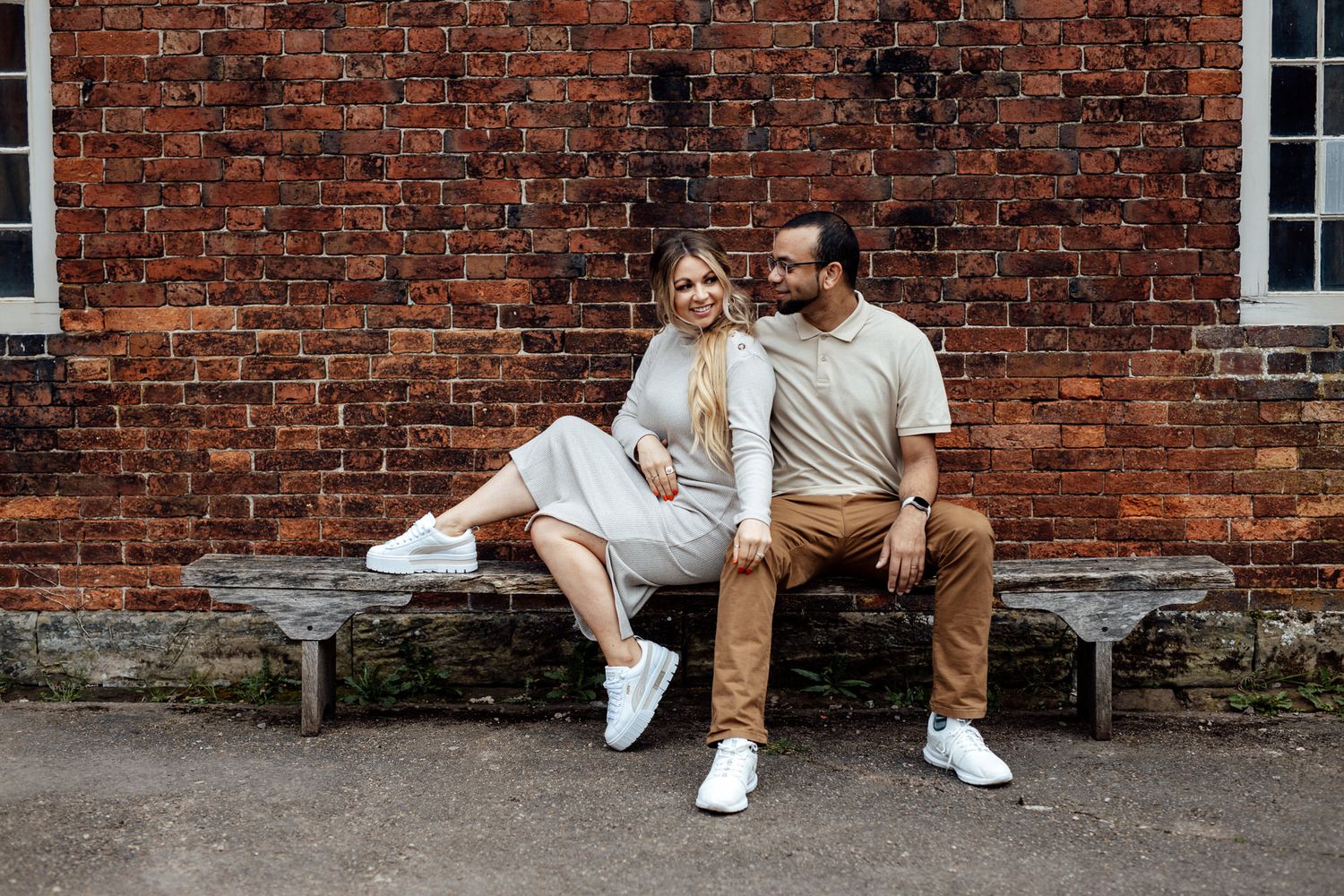 A couple poses playfully against a rustic brick wall in coordinated white and brown outfits.
