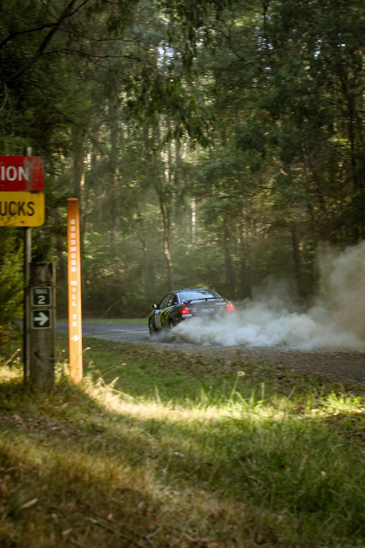 Rally car kicking up dust while racing past directional signs on a wooded course.