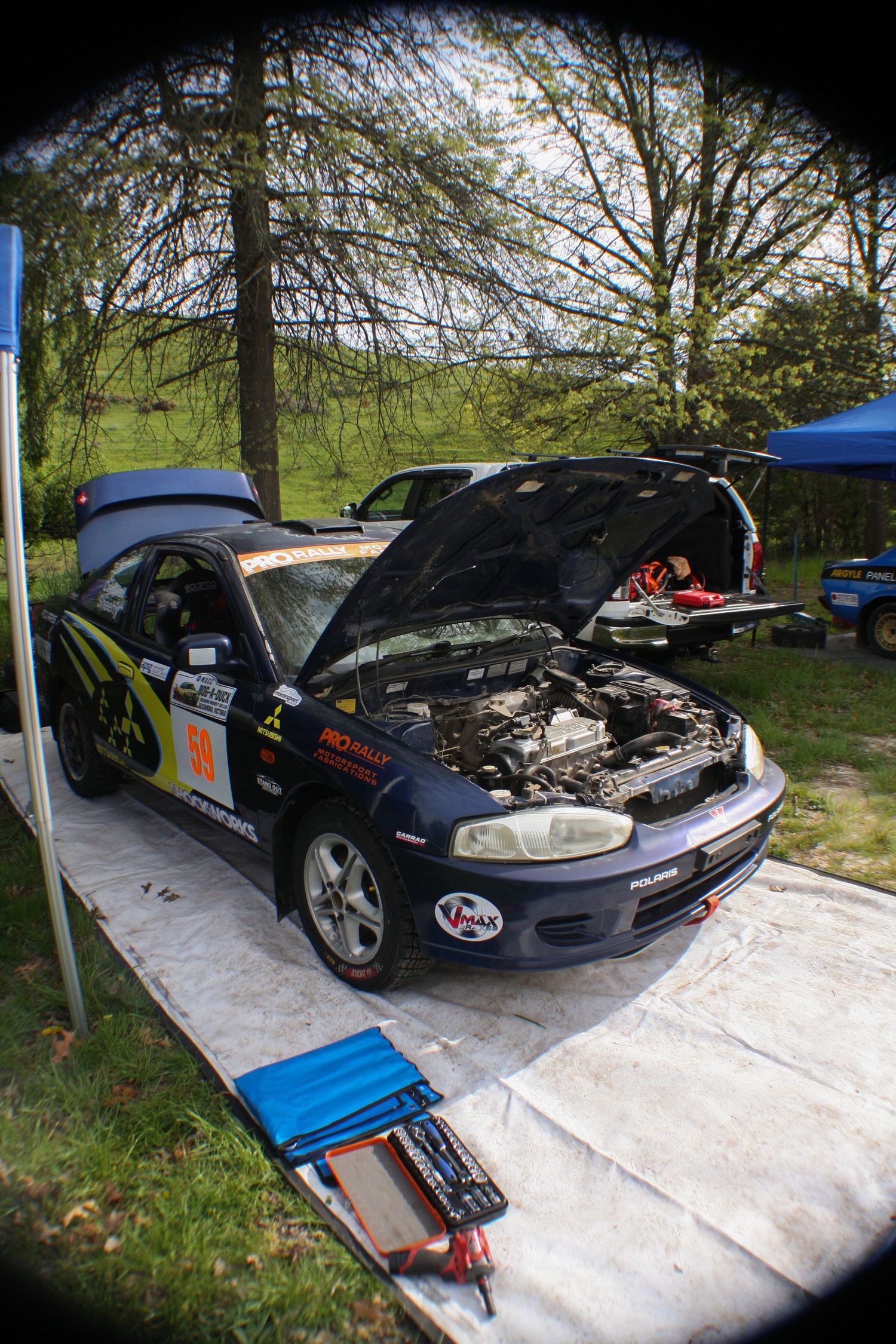 Car hood open showing engine and mechanical components under a blue tent at an outdoor event.