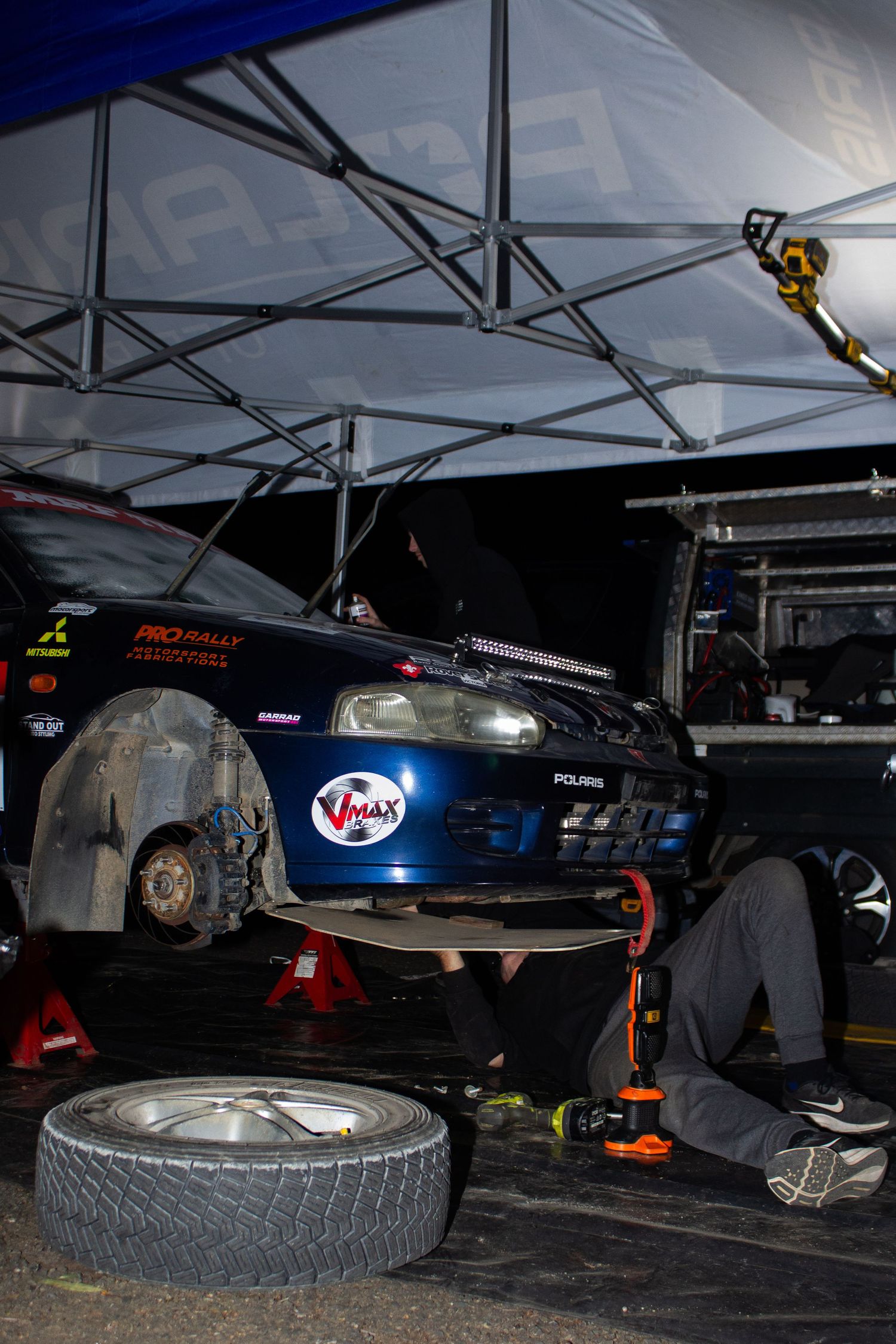 Rally car undergoing maintenance in pit area with exposed wheel and mechanics working underneath.
