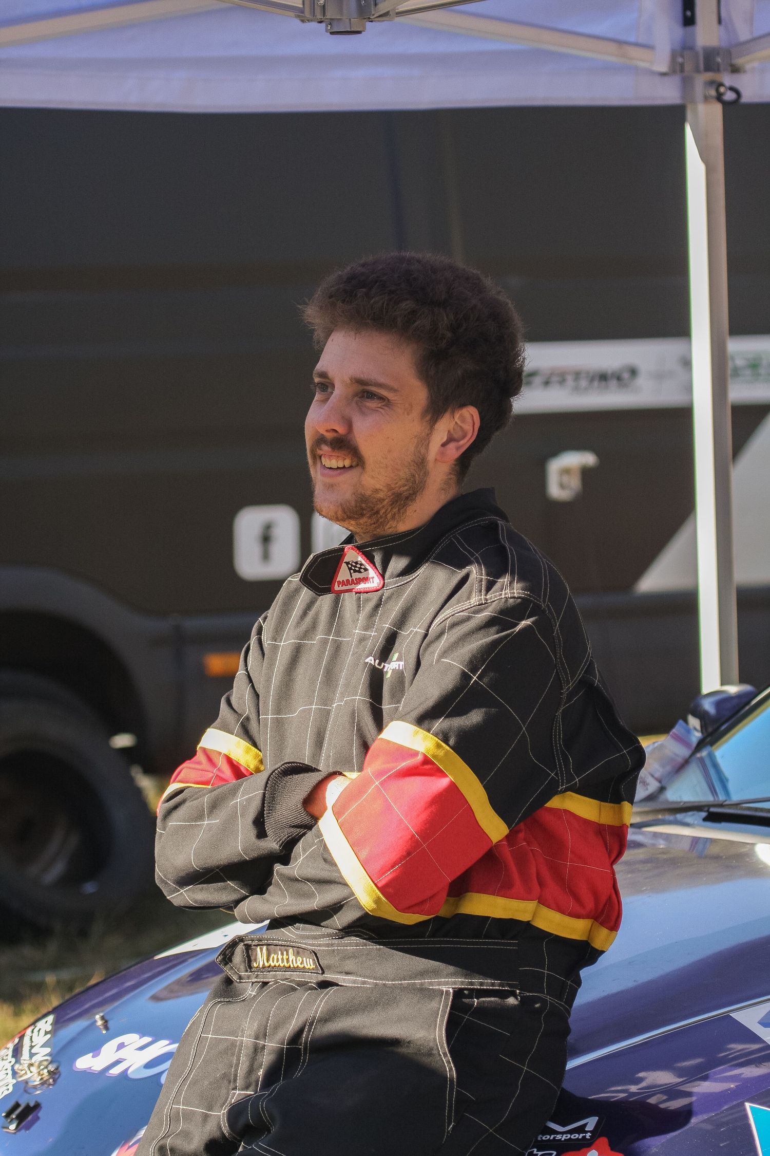 Racing team members in uniform relaxing beside a blue car at an outdoor event.