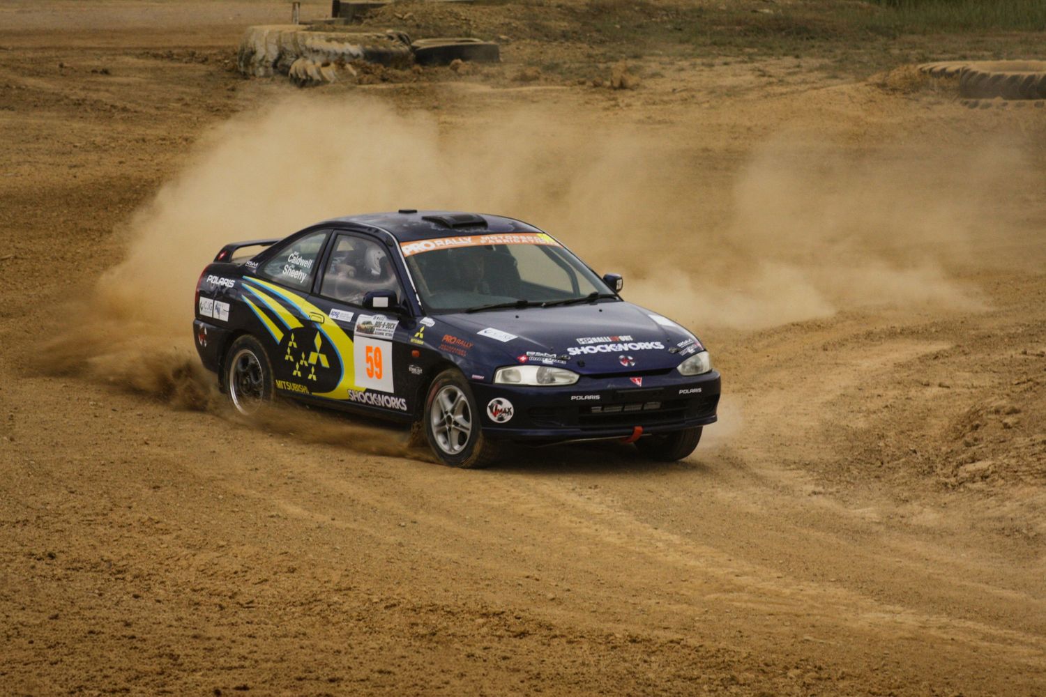 Dark colored rally car sliding through dirt corner during race.