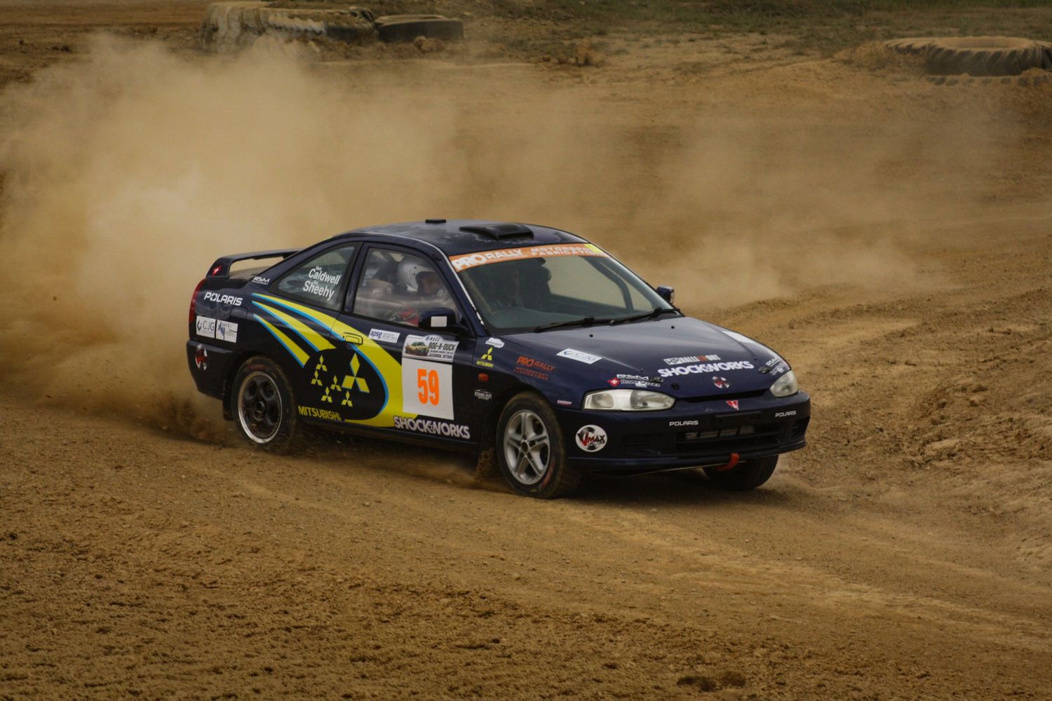 Rally cars racing in formation through dusty dirt course corner.