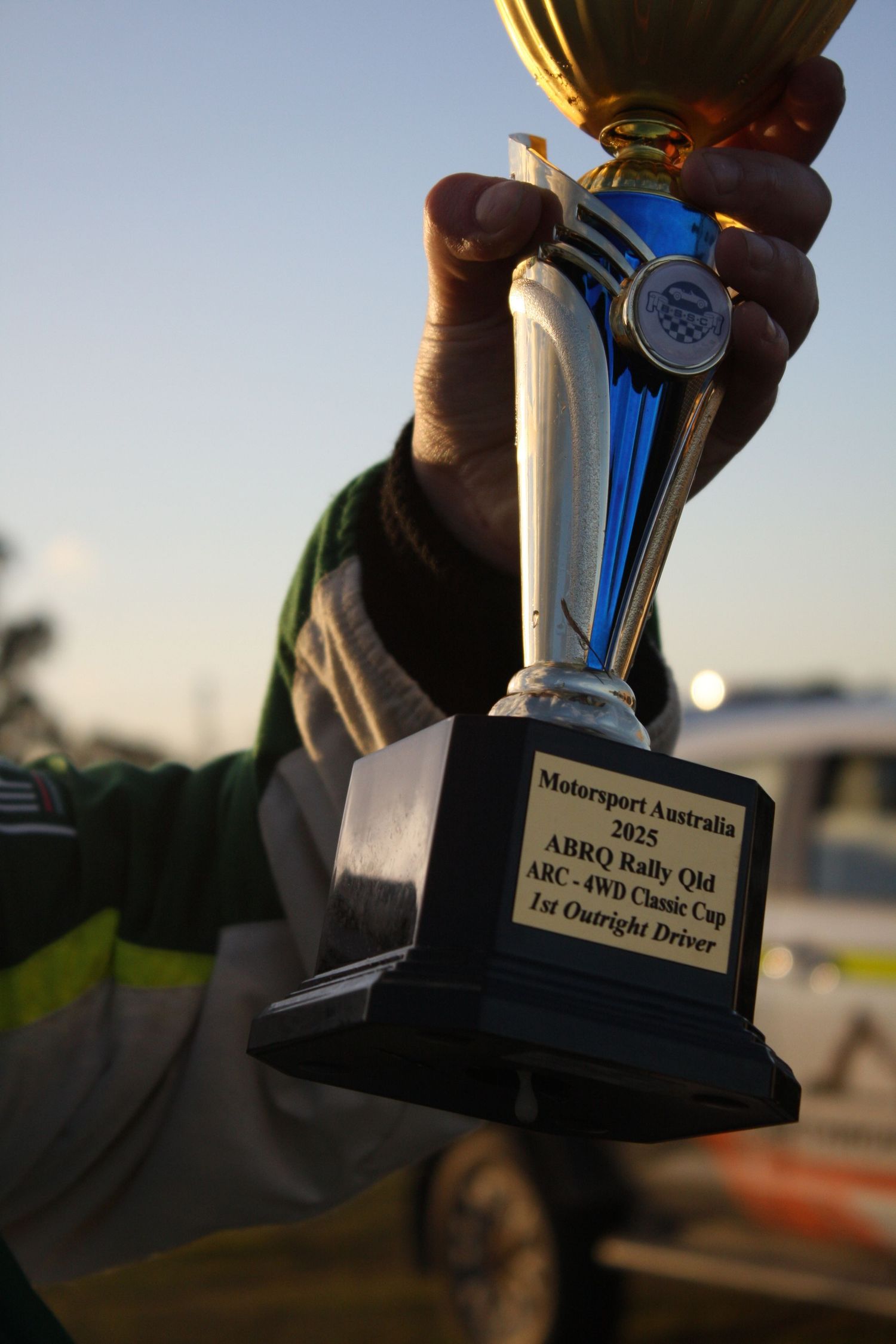 A hand holds up a motorsport trophy against a sunset sky.
